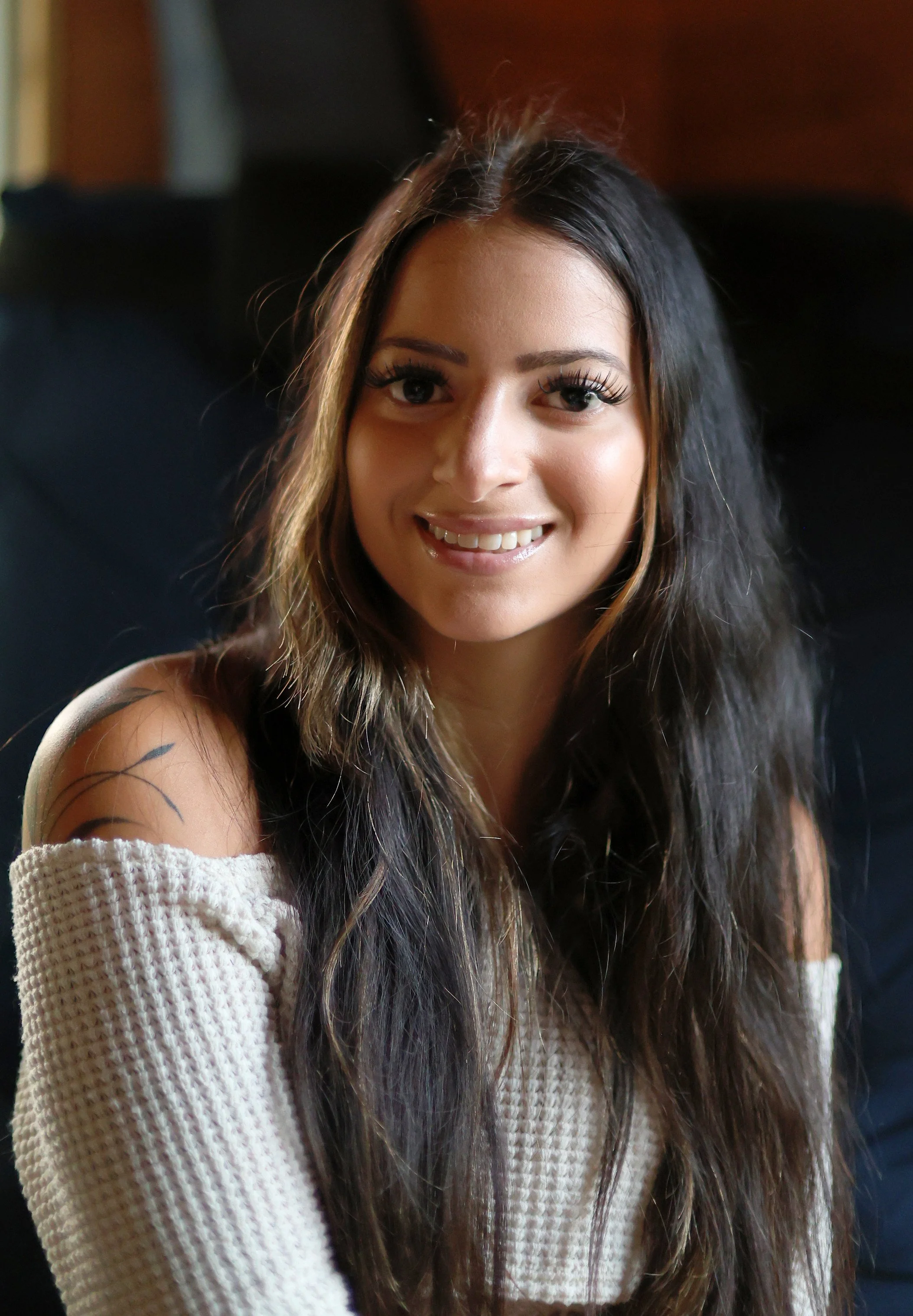 Close-up of a young woman with long dark hair smiling at the camera, wearing a cream-colored, off-the-shoulder sweater.