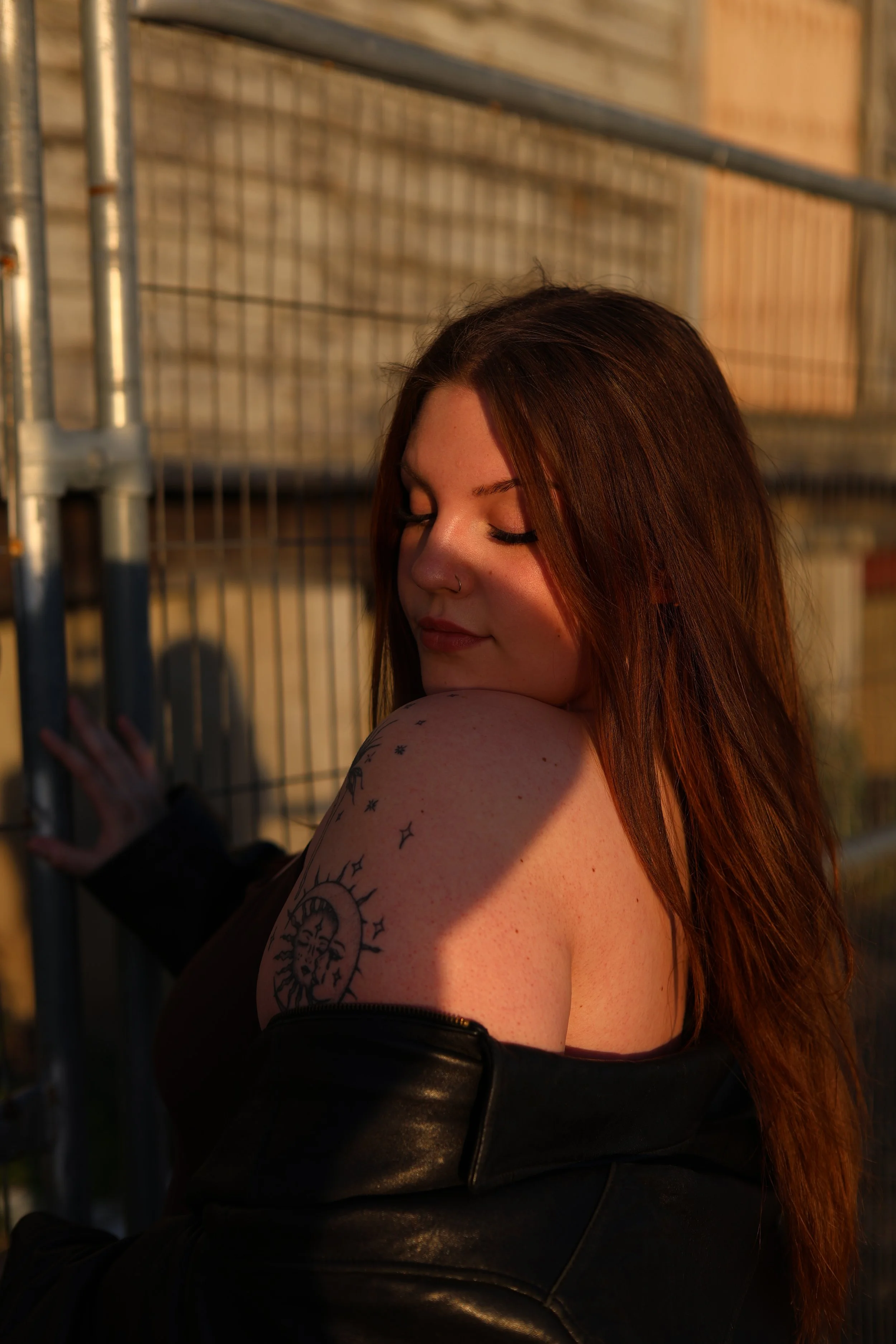 A woman with long reddish-brown hair and tattoos on her shoulder is leaning against a metal fence, with her eyes closed, in warm golden lighting.