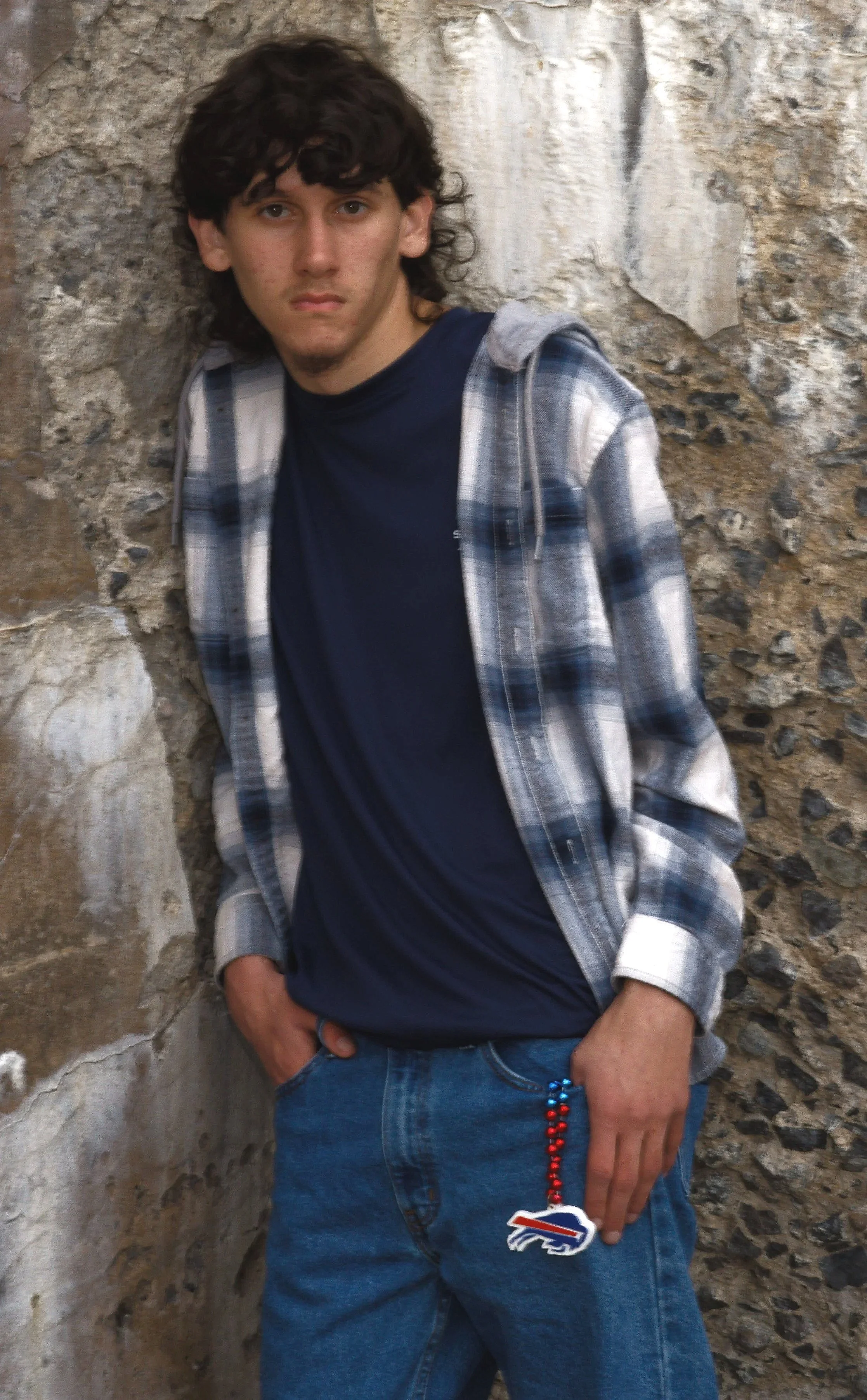 A young man with dark curly hair wearing a navy shirt, plaid jacket, and jeans, standing against a stone wall with one hand in his pocket.