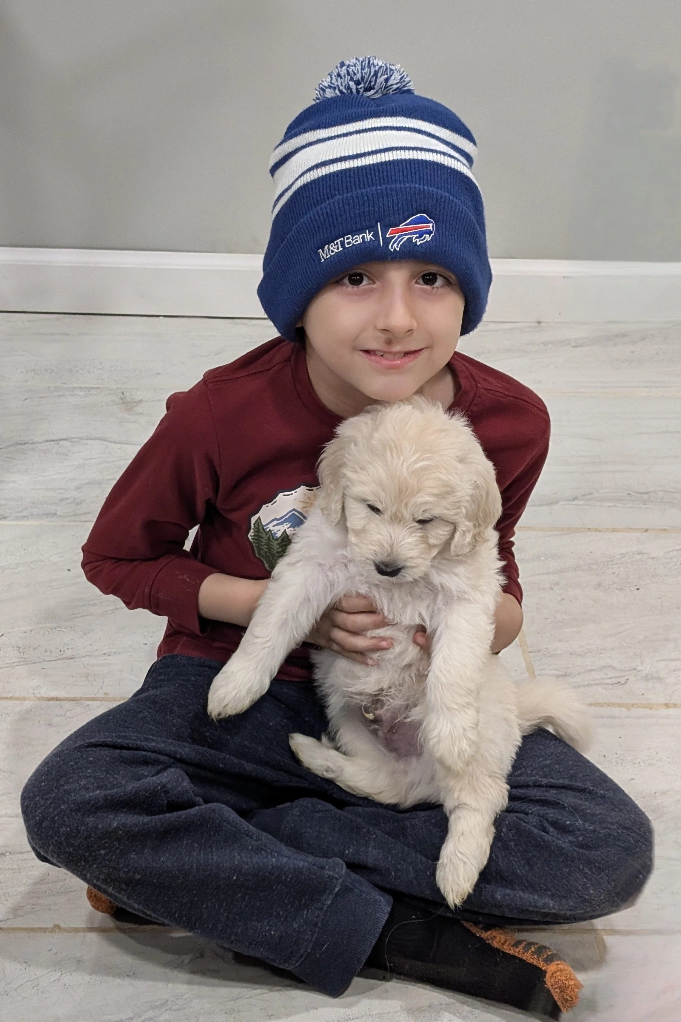 A young boy sitting cross-legged on the floor holding a fluffy yellow puppy, wearing a blue winter hat with white stripes and pom-pom, a maroon shirt, and dark blue jeans.