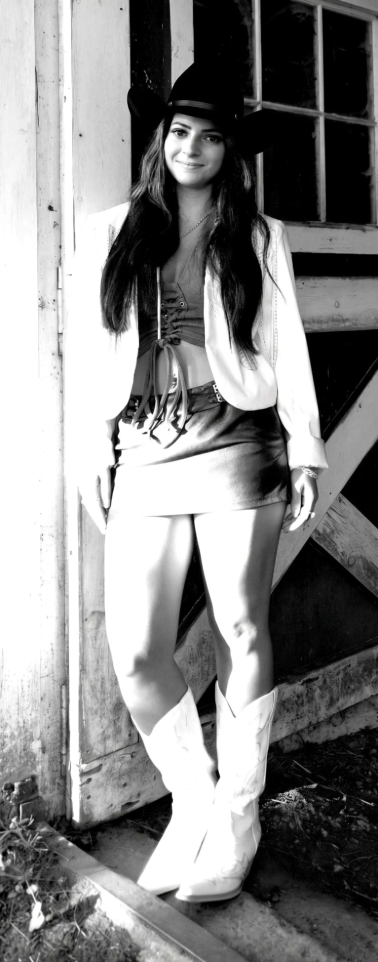 A young woman wearing a cowboy hat, a blazer, a crop top, a leather skirt, and cowboy boots standing indoors near a rustic wooden wall and window.