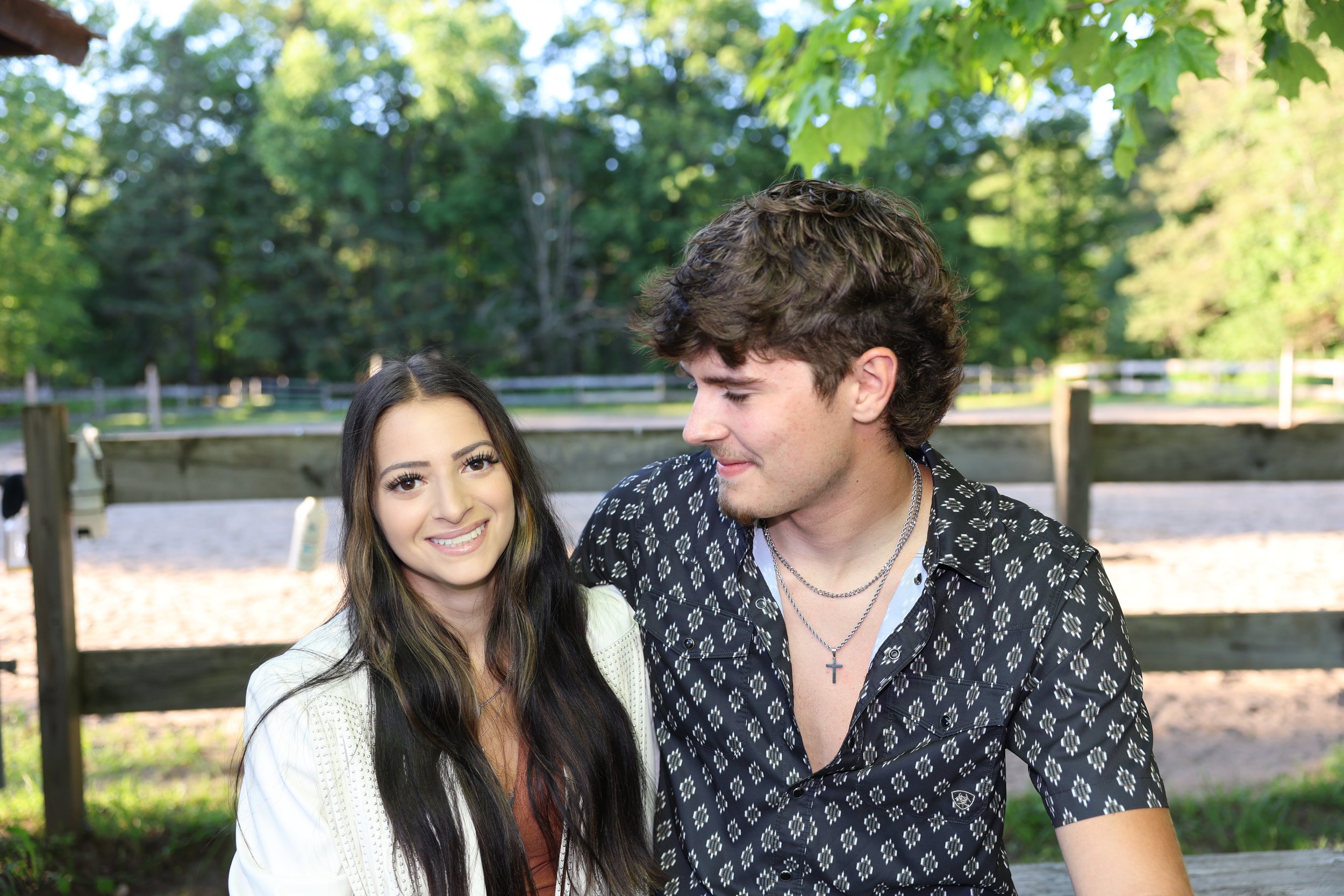 A young woman with long, dark hair and a young man with curly brown hair, wearing a patterned shirt and necklaces, are outdoors in a park with green trees and a wooden fence in the background. They are standing close together and smiling.