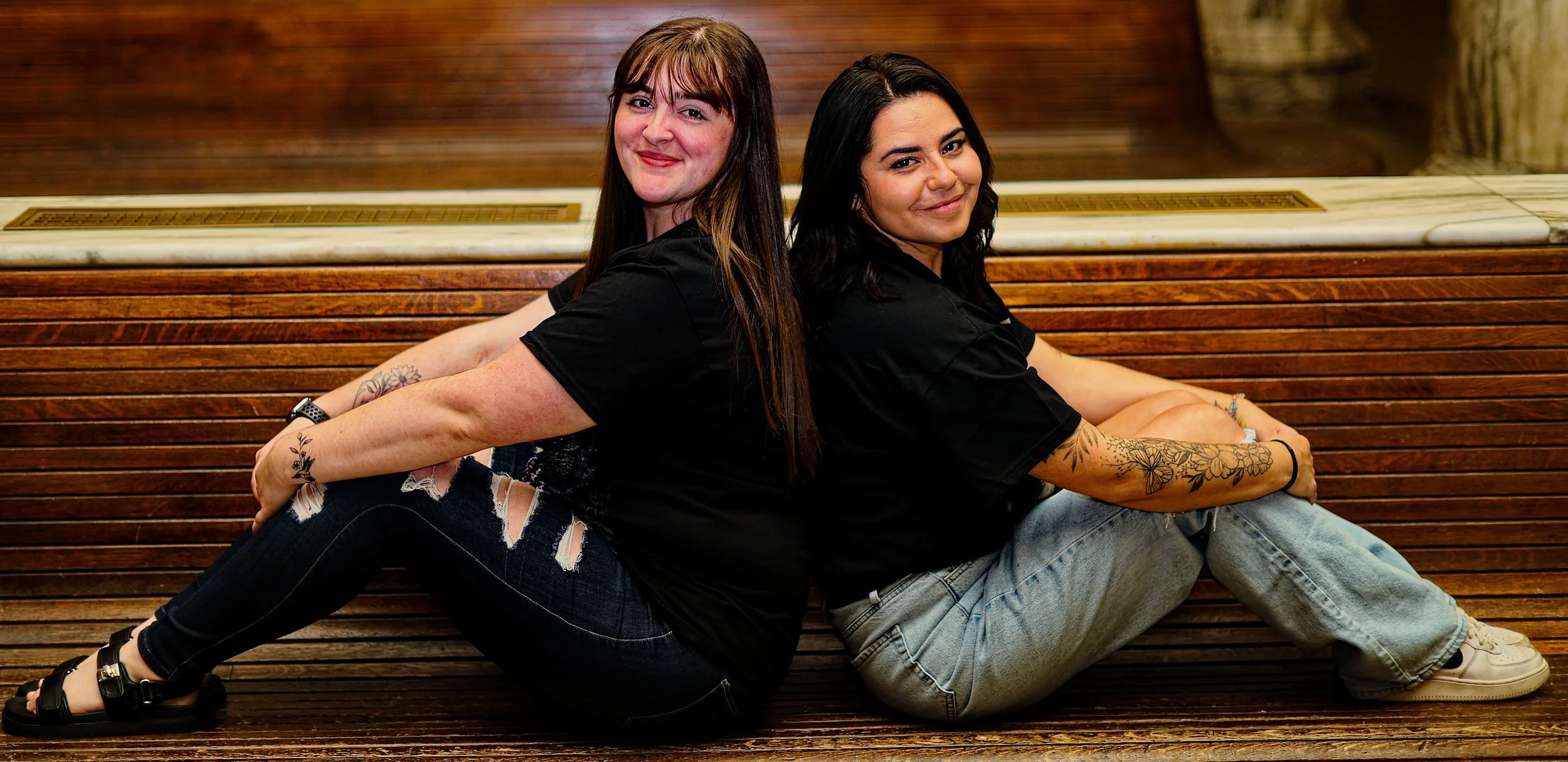 Two women sitting back-to-back on a wooden bench, smiling and looking at the camera.