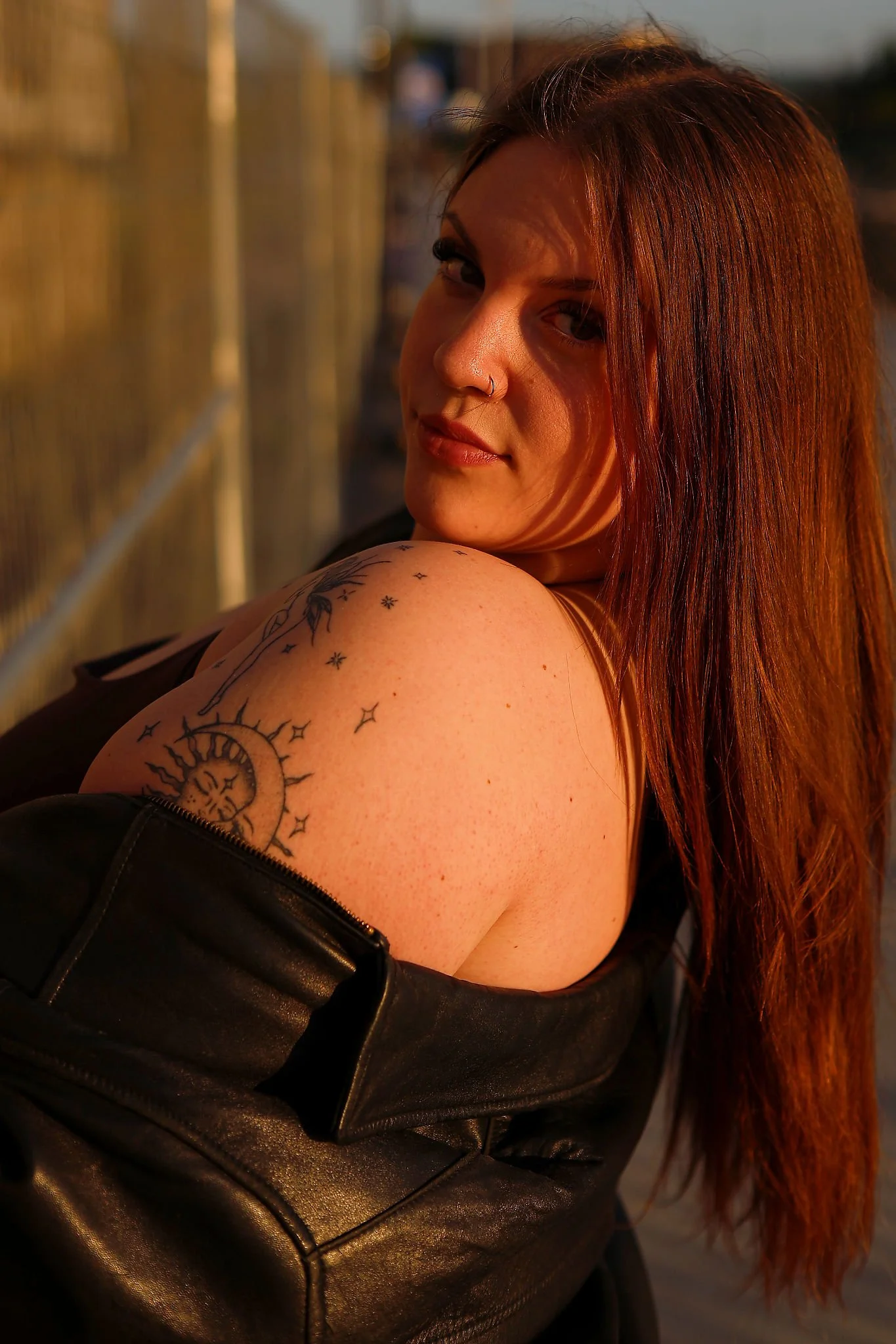 A woman with long reddish hair looking over her shoulder at the camera, wearing a black leather jacket with visible tattoos on her shoulder, against a blurred background with warm sunlight.