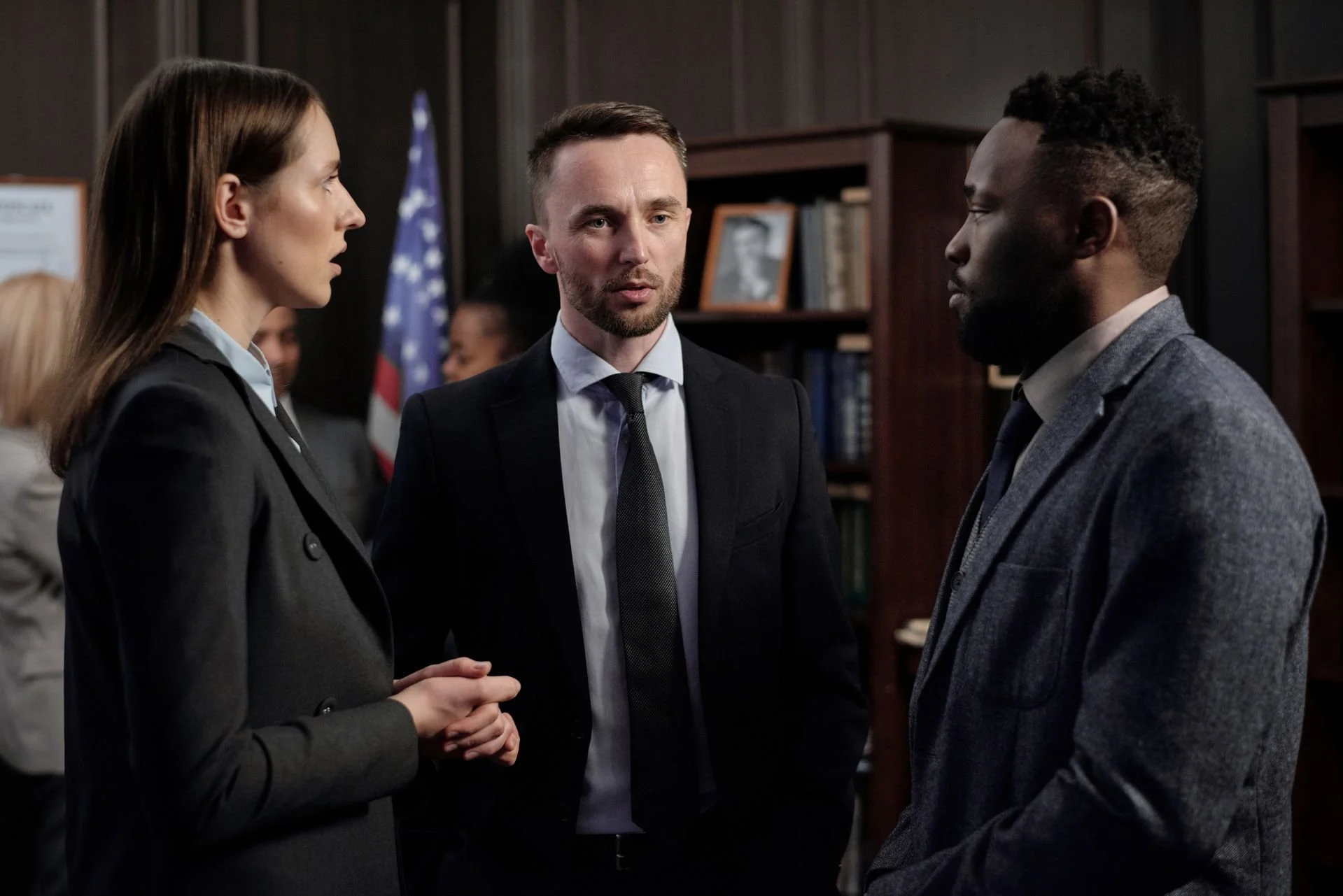 Three professionals, two men and one woman, engaged in a serious conversation in an office setting, with bookshelves and an American flag in the background.