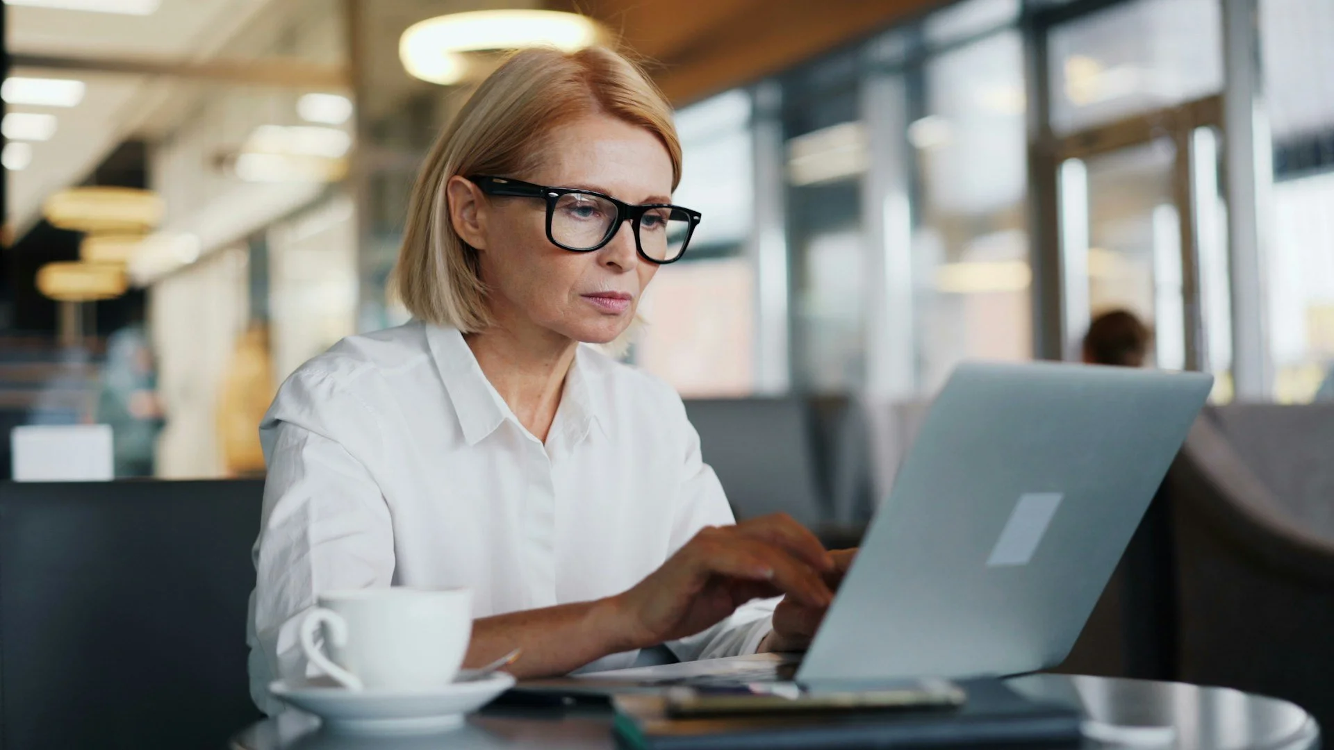 A middle-aged woman with blonde hair, wearing a white blouse and black glasses, working on a silver laptop in a modern, well-lit café or coffee shop.