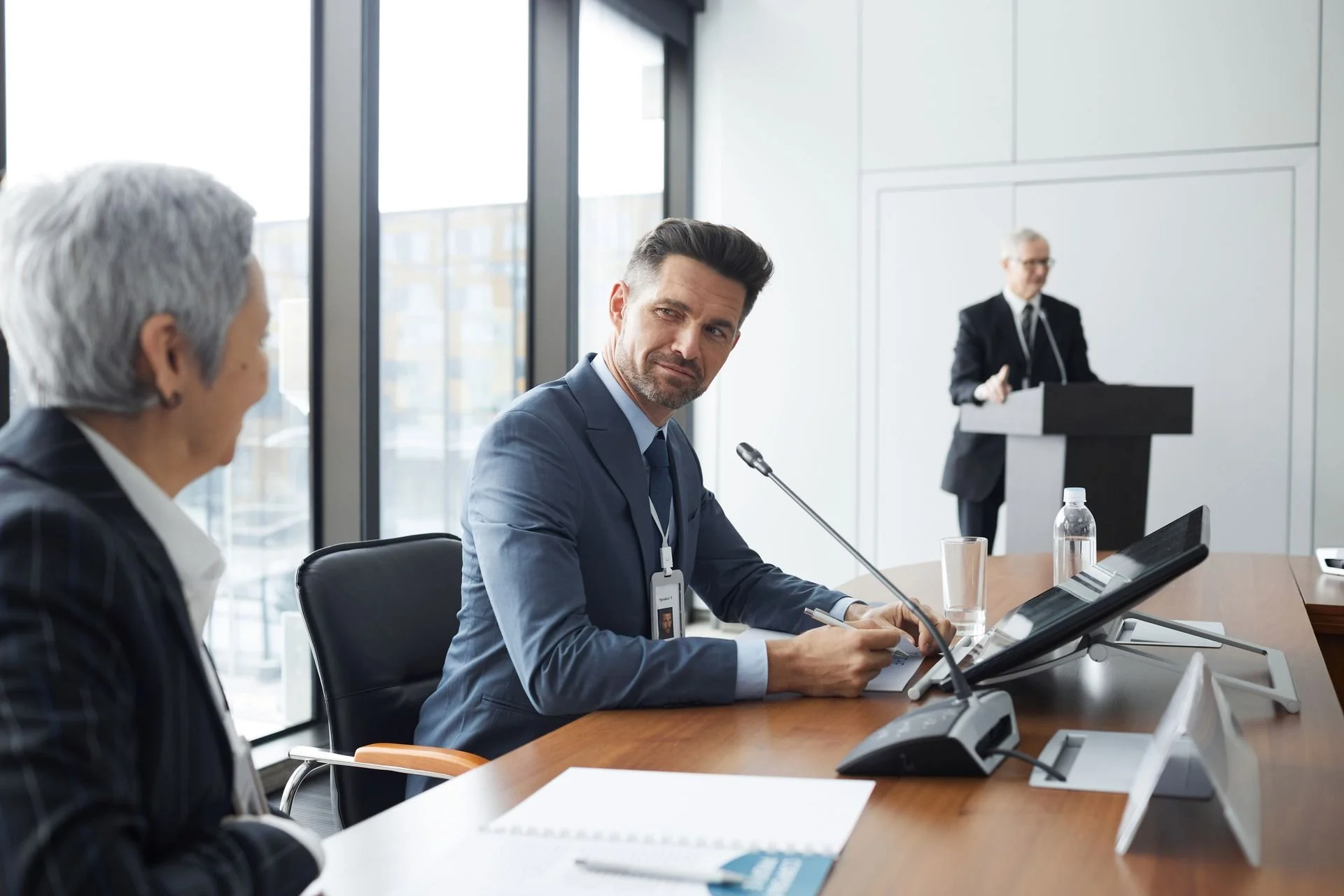 Business meeting in a conference room with three men engaged in discussion. One man at the table is holding a pen and wearing an ID badge, while another is speaking at a podium near the wall.