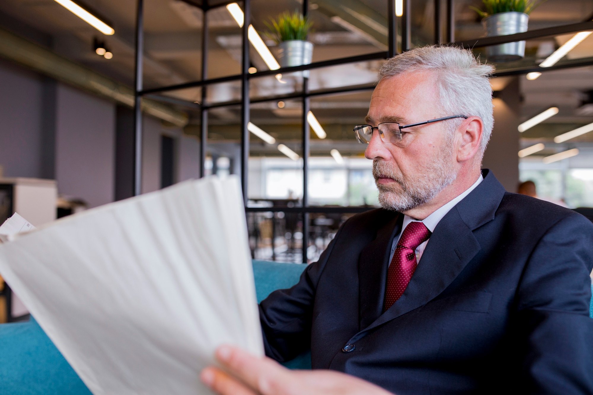 An older man with gray hair, glasses, and a beard dressed in a dark suit and red tie, sitting in a modern office or café, looking intently at a large folder or document he is holding.