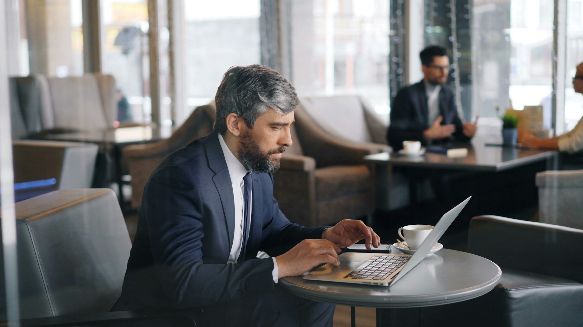 A middle-aged man with gray hair and beard, wearing a dark suit, sitting at a café table working on a laptop, with a coffee cup in front of him.