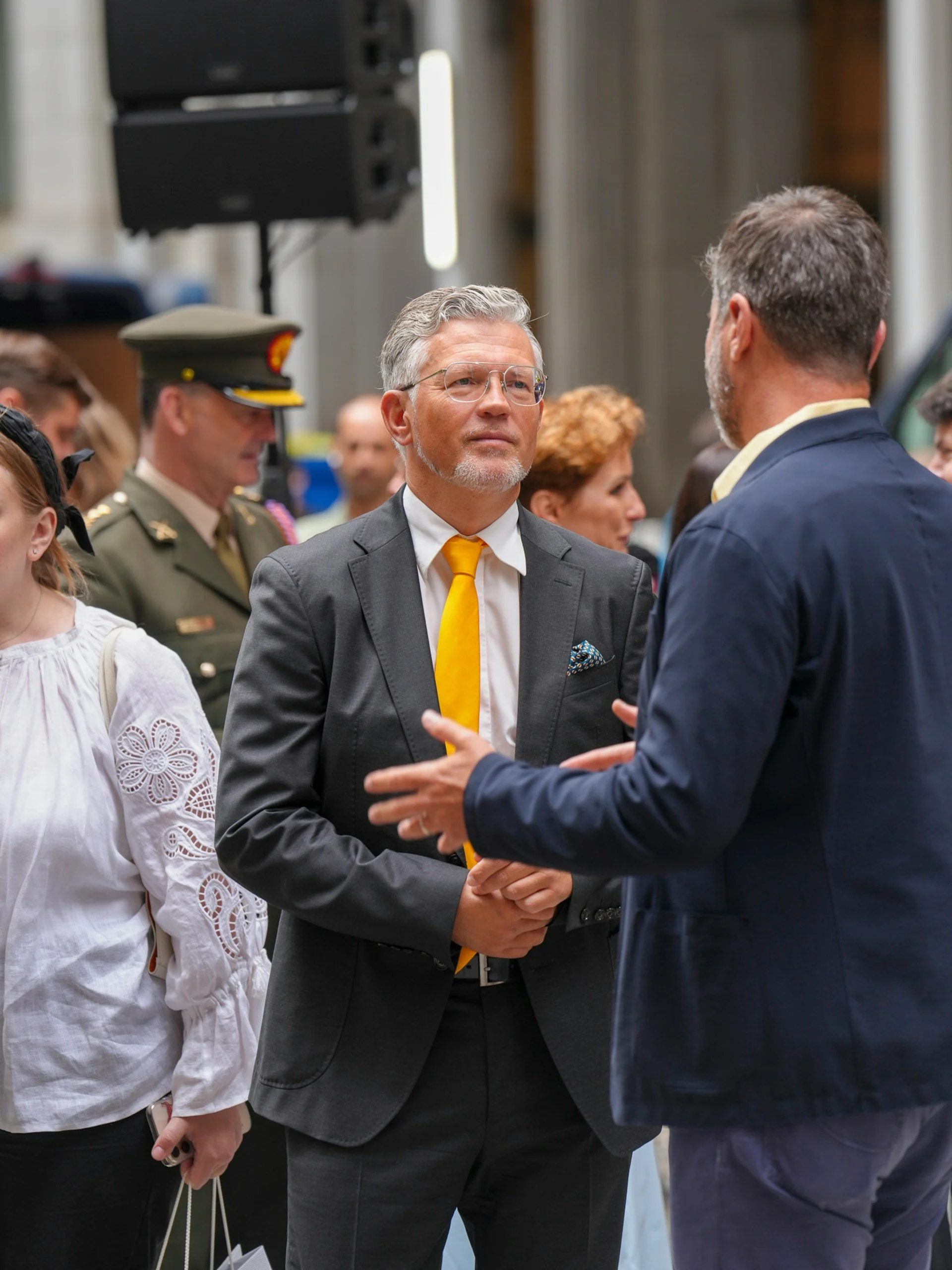 People engaging in conversation at an indoor event, including a man in a grey suit with glasses and a yellow tie, and a man in a blue jacket. There are other people in the background.