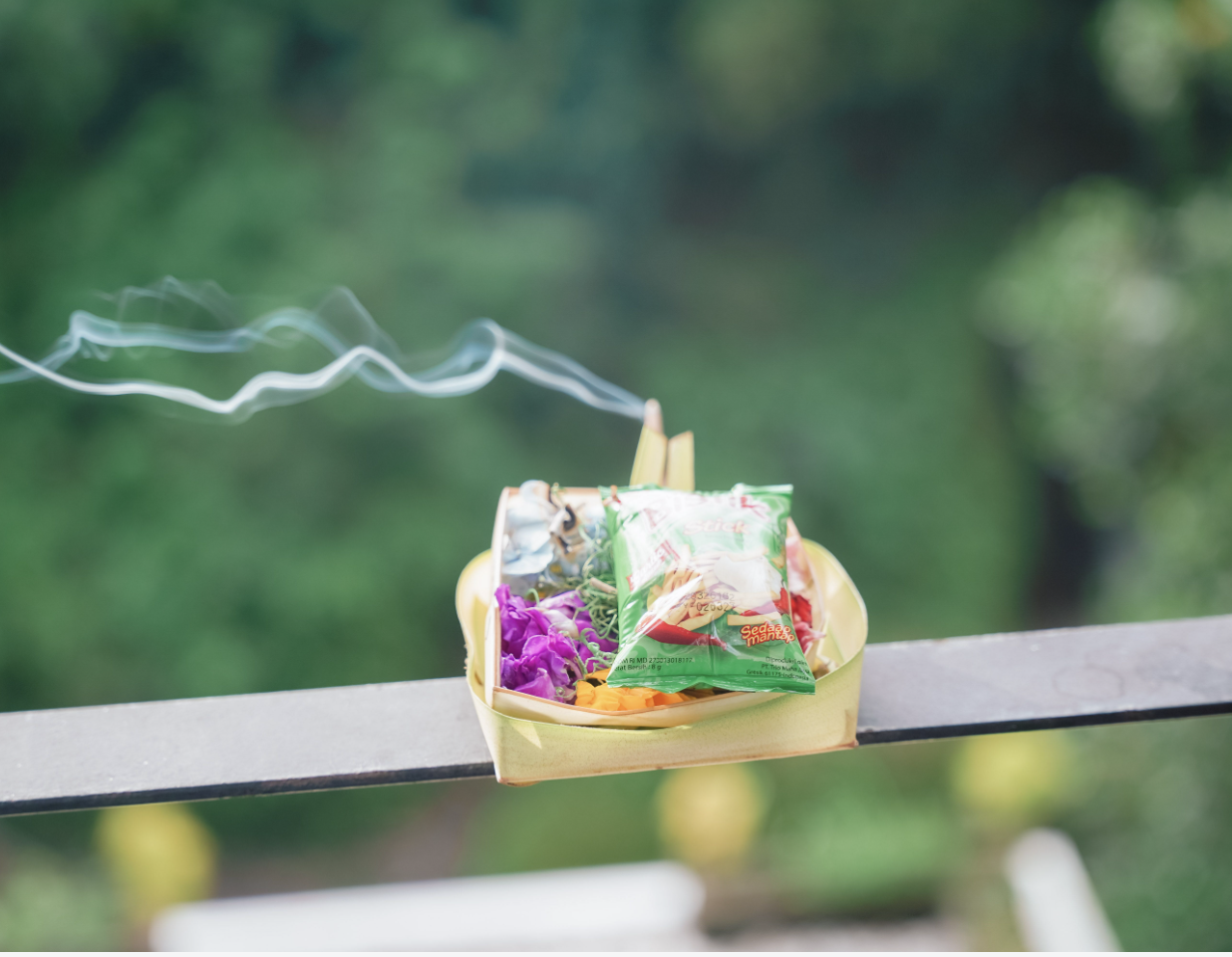A small yellow tray on a metal railing containing snacks and a lit cigarette, with smoke curling into the air and a blurred green background.
