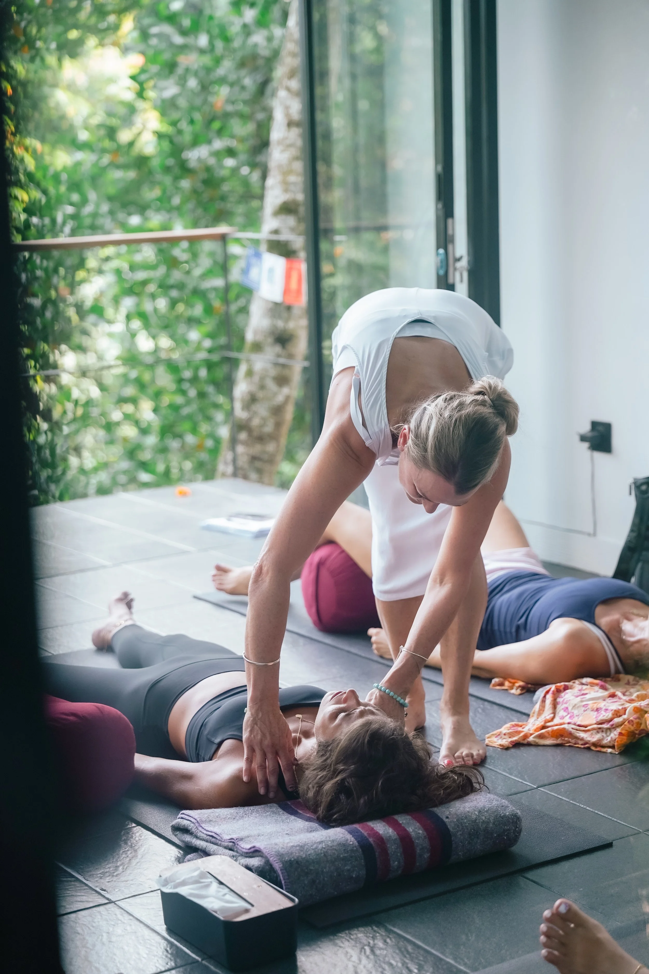A woman assisting another woman in a yoga pose during a yoga class in a bright room with large windows and lush green trees outside.