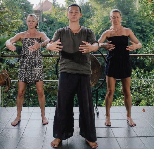 Three people practicing yoga outdoors on a deck with jungle foliage background, two women and one man, standing with arms extended and palms facing down.