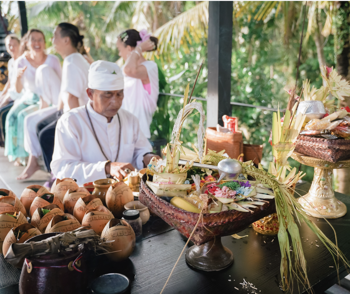 People dressed in white traditional clothing participate in a religious or cultural ceremony outdoors, surrounded by lush green foliage, with offerings and decorative items on a table.