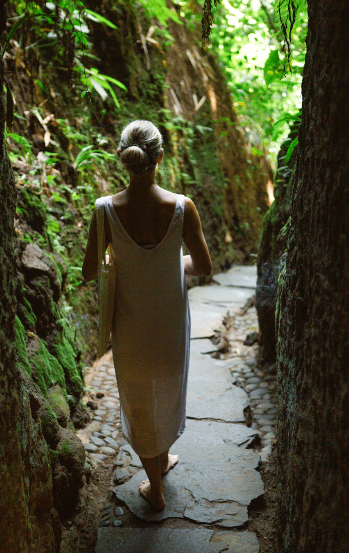 A woman with gray hair in a bun walking barefoot on a narrow stone path through a green jungle.