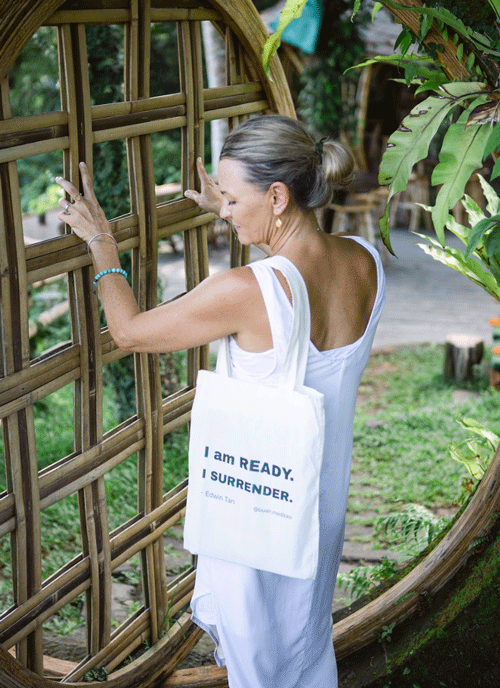 An elderly woman with gray hair tied in a bun, wearing a white sleeveless dress, stands outside near lush green foliage and a bamboo structure. She is holding the bamboo with her left hand and adjusting or examining something on it with her right hand. She carries a white tote bag with the text 'I AM READY. I SURRENDER.' printed on it.