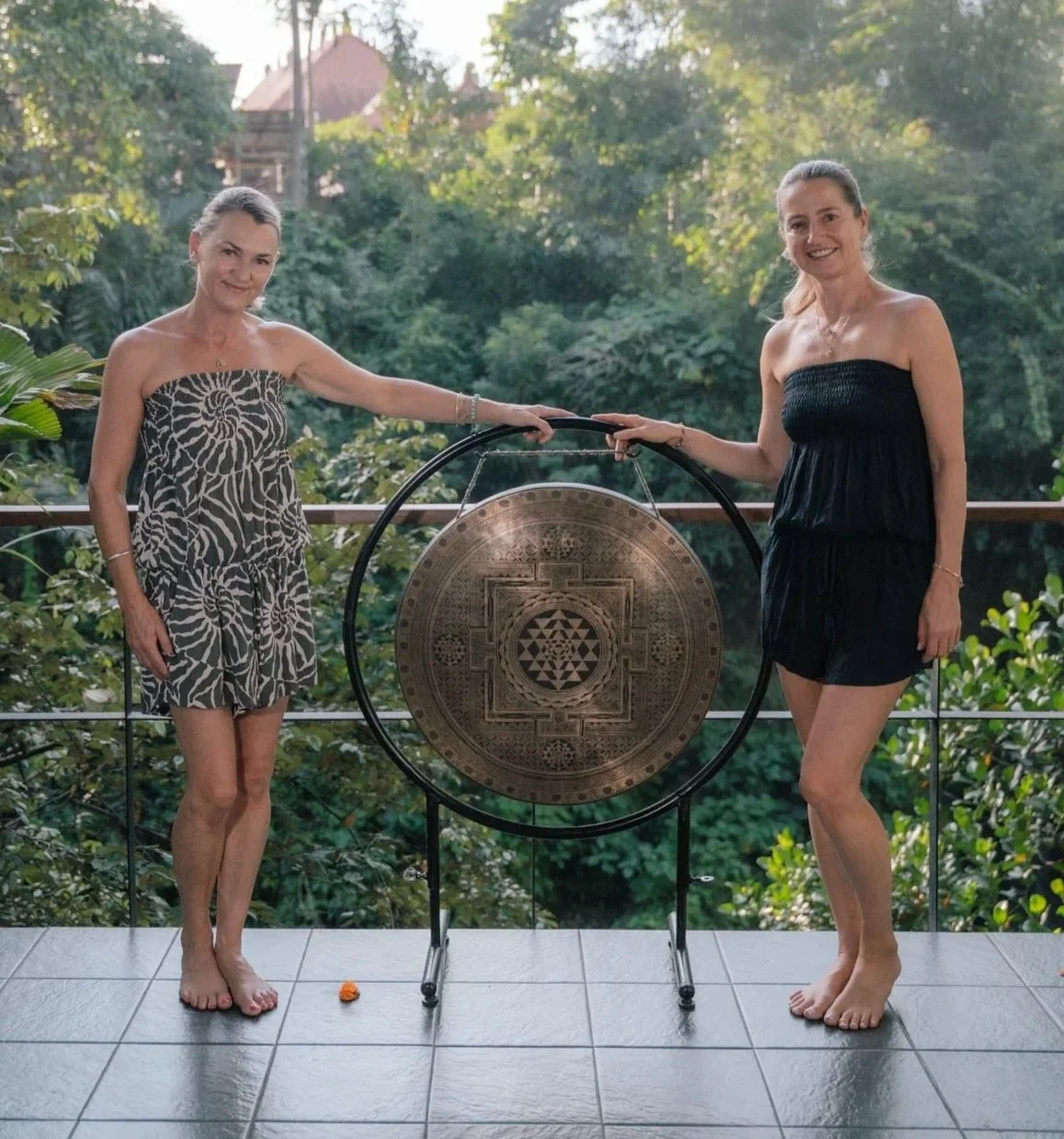 Two women in strapless dresses standing outdoors on a balcony with greenery in the background, holding a large decorative gong.