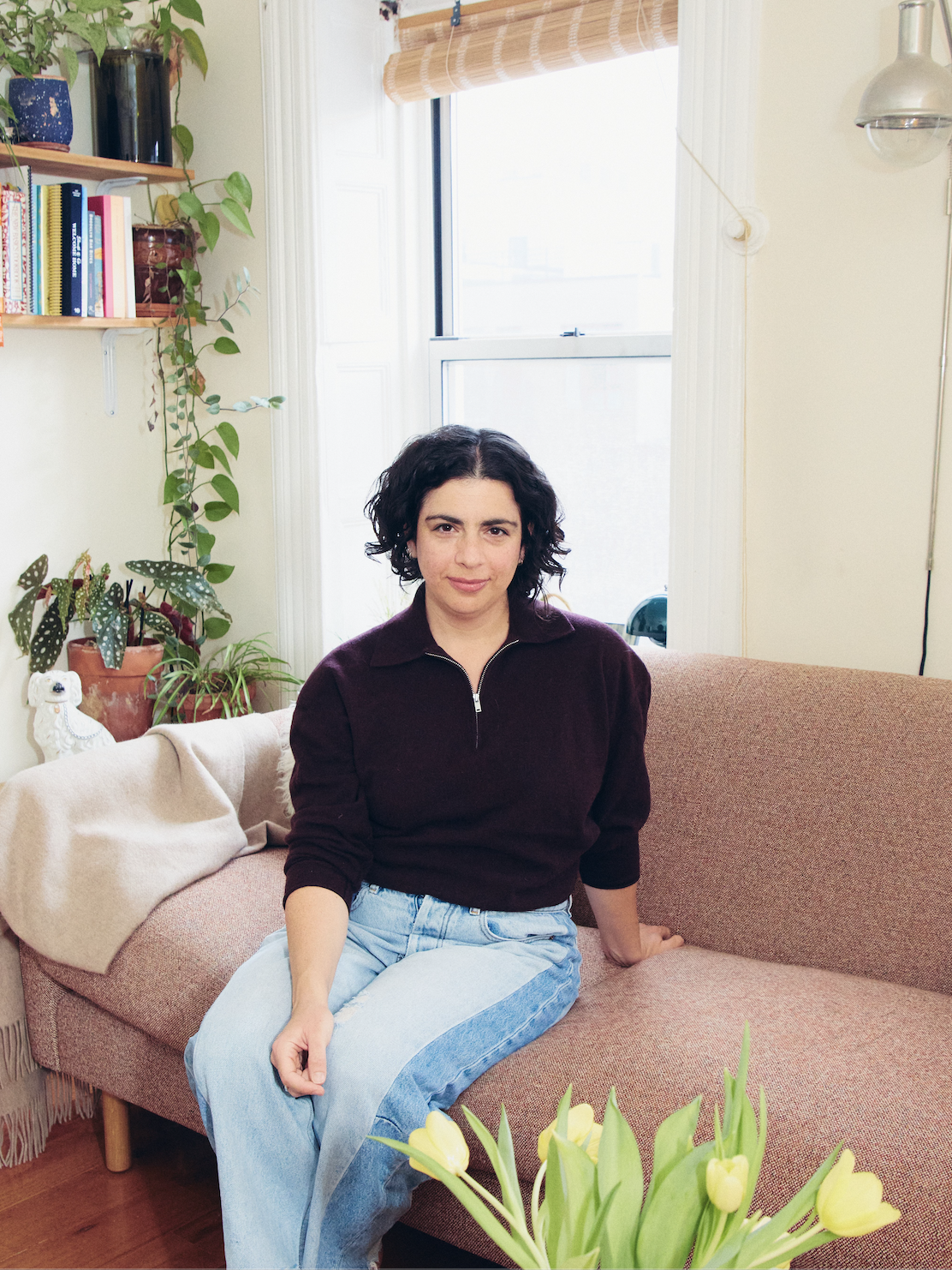 A woman with dark curly hair sitting on a pinkish sofa in a cozy living room. She is wearing a dark purple zip-up sweater and light blue jeans, looking at the camera with a slight smile. Behind her are green plants, a window with a bamboo shade, and a wooden bookshelf with various books and decorative items. In the foreground, there is a vase of yellow tulips.