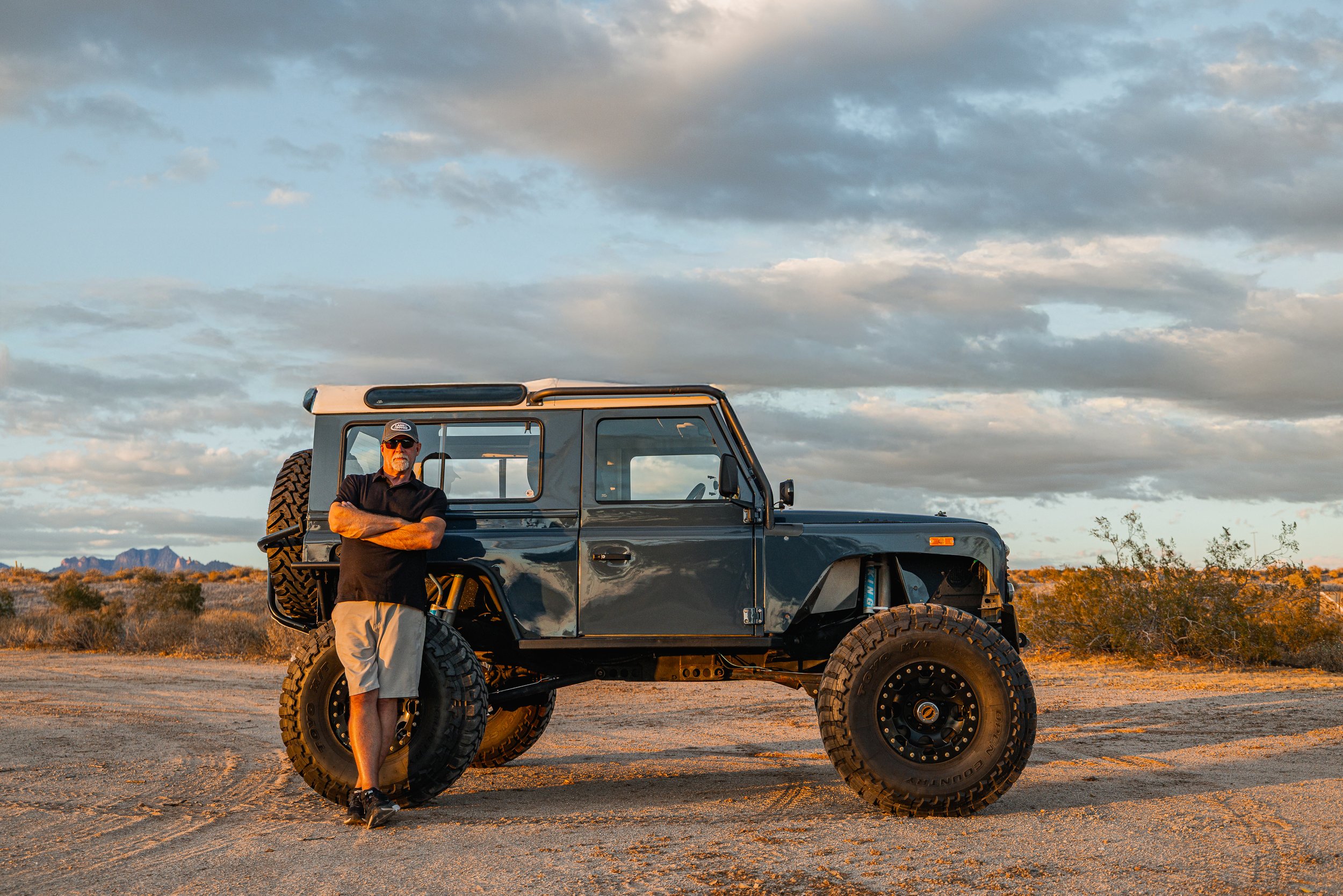 A man standing with arms crossed in front of a black off-road vehicle in a desert landscape during sunset, with mountains in the background and cloudy sky.