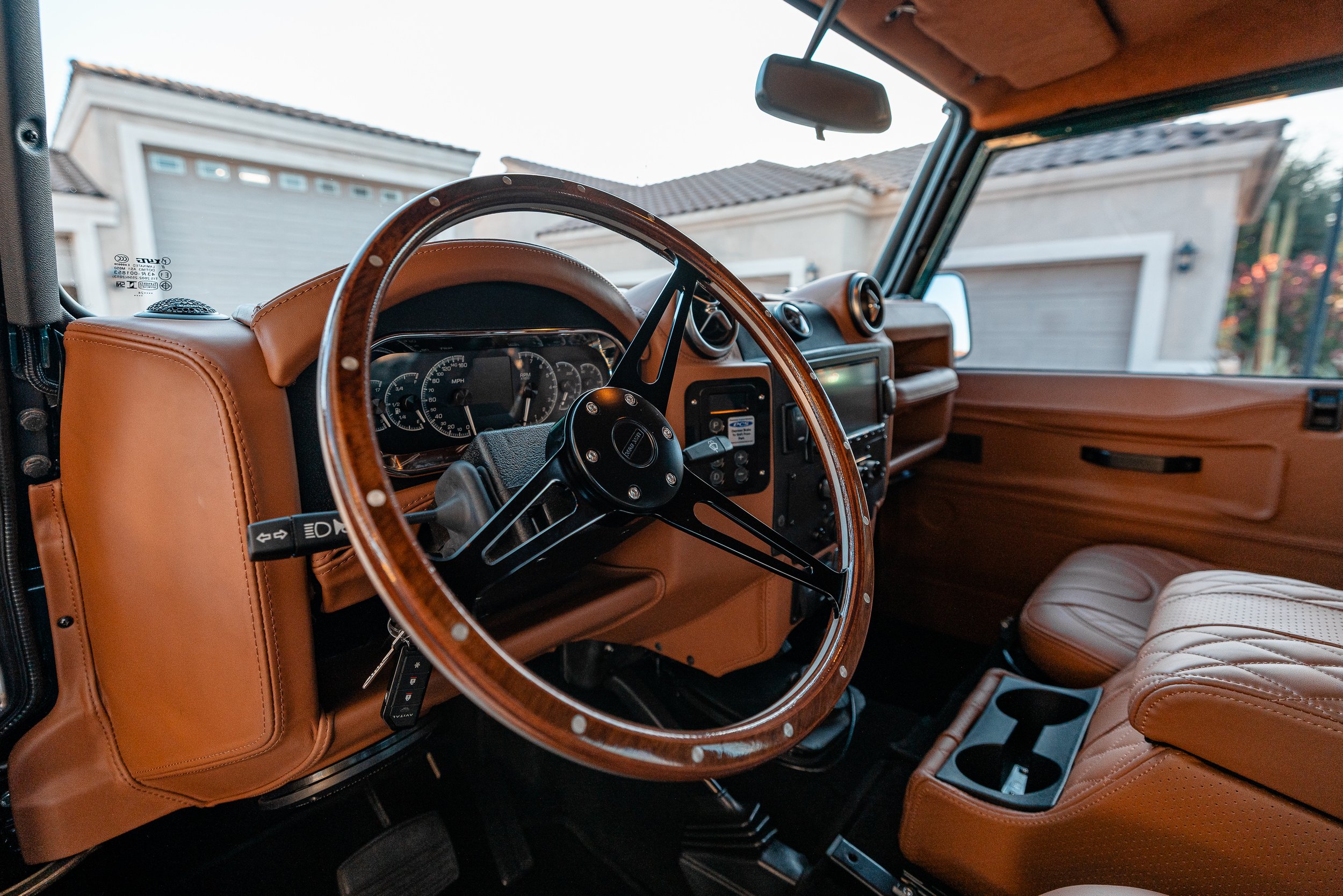 Interior of a vintage car with brown leather seats and a classic steering wheel, parked in a residential driveway during daytime.