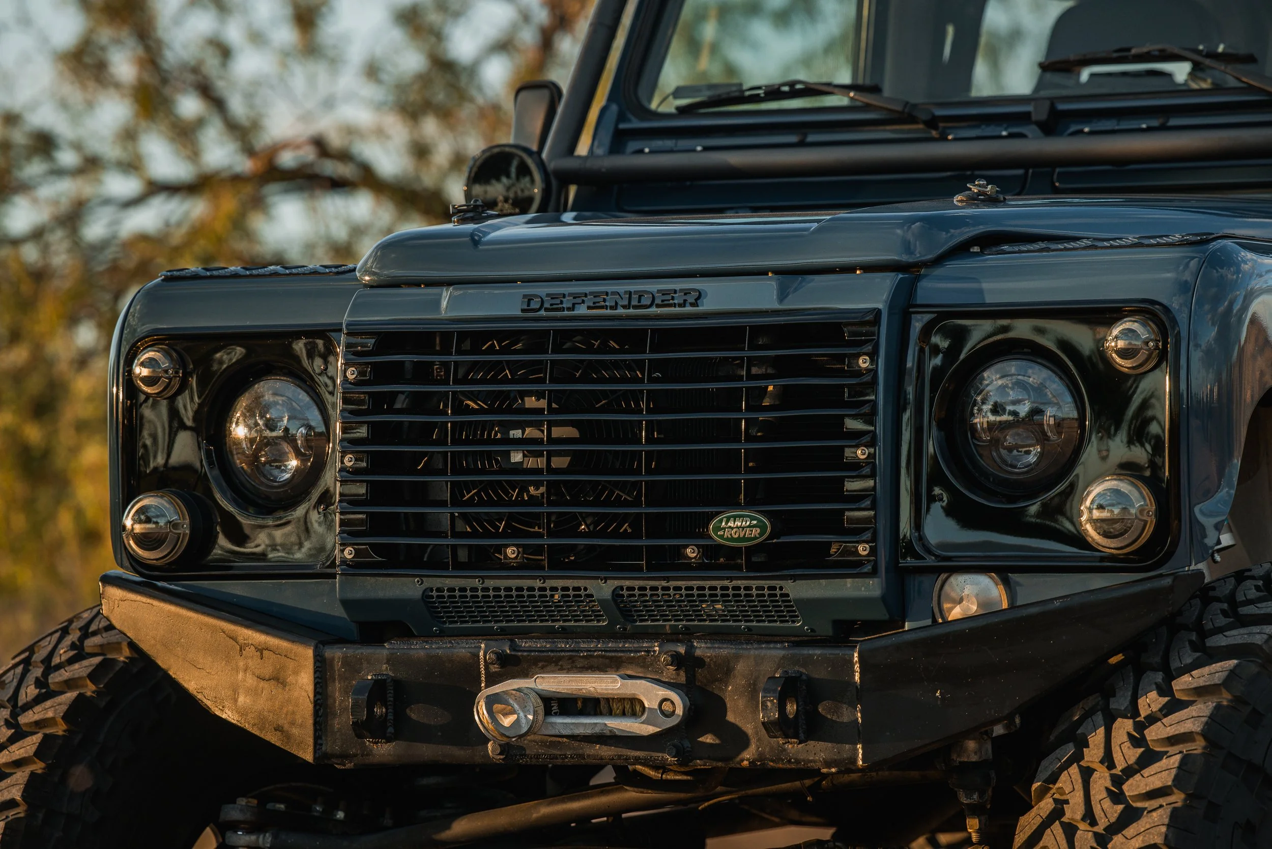 Close-up of the front of a black Land Rover Defender off-road vehicle, featuring its grille, headlights, and brand badge, with a blurred outdoor background.