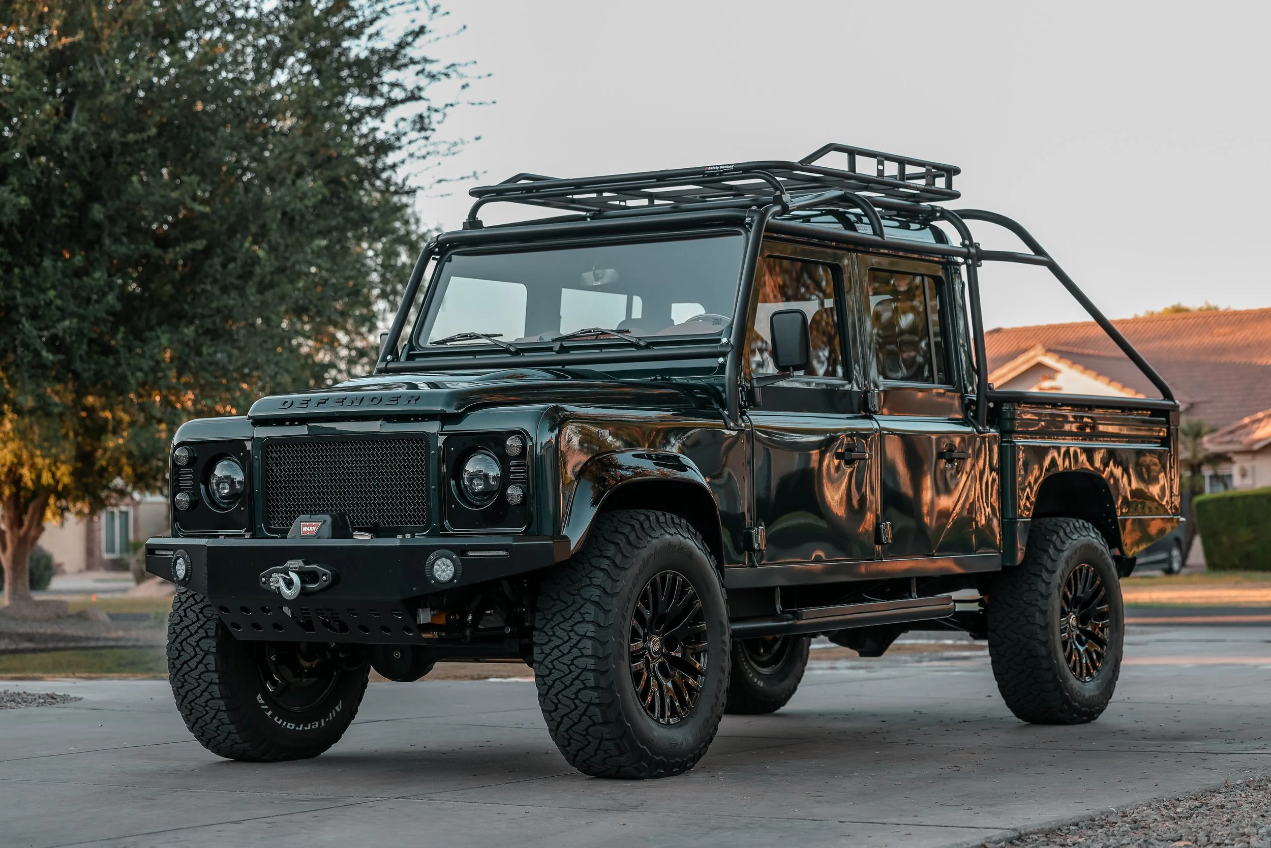 Black Land Rover Defender pickup truck parked outdoors during sunset, with trees, a house, and a clear sky in the background.