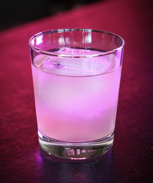 A clear glass filled with a pink drink and ice cubes, placed on a dark pink surface.