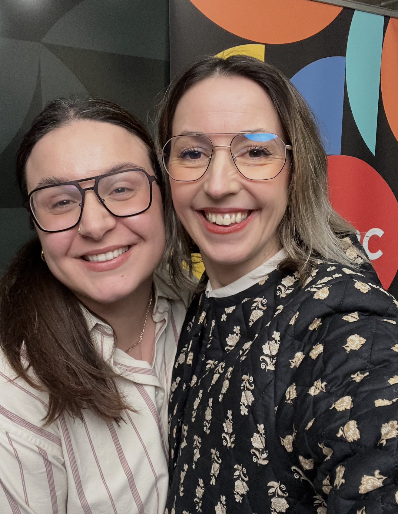 Claire Bonnyman and JoAnne Pearce smiling and taking a selfie in front of a colorful geometric background.