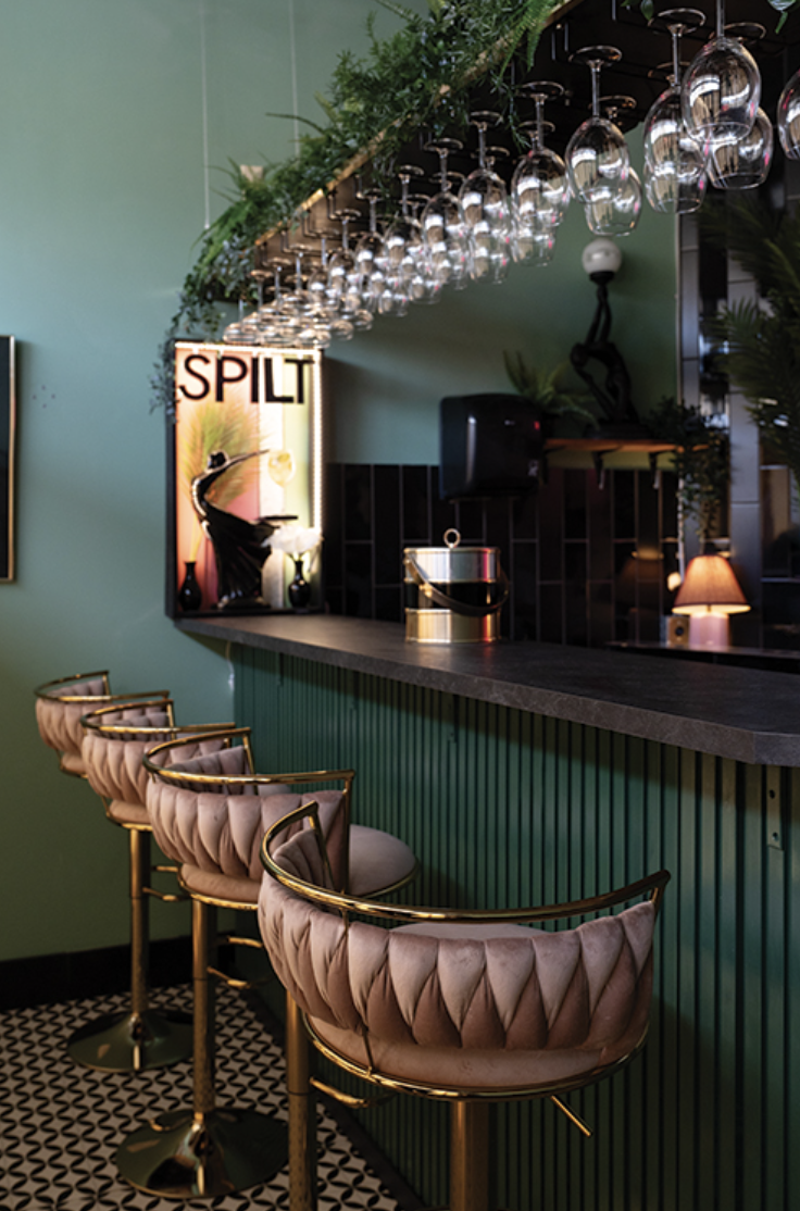 A bar with gold and pink upholstered bar stools, hanging wine glasses, and tropical plants, in a stylish nightclub or lounge.