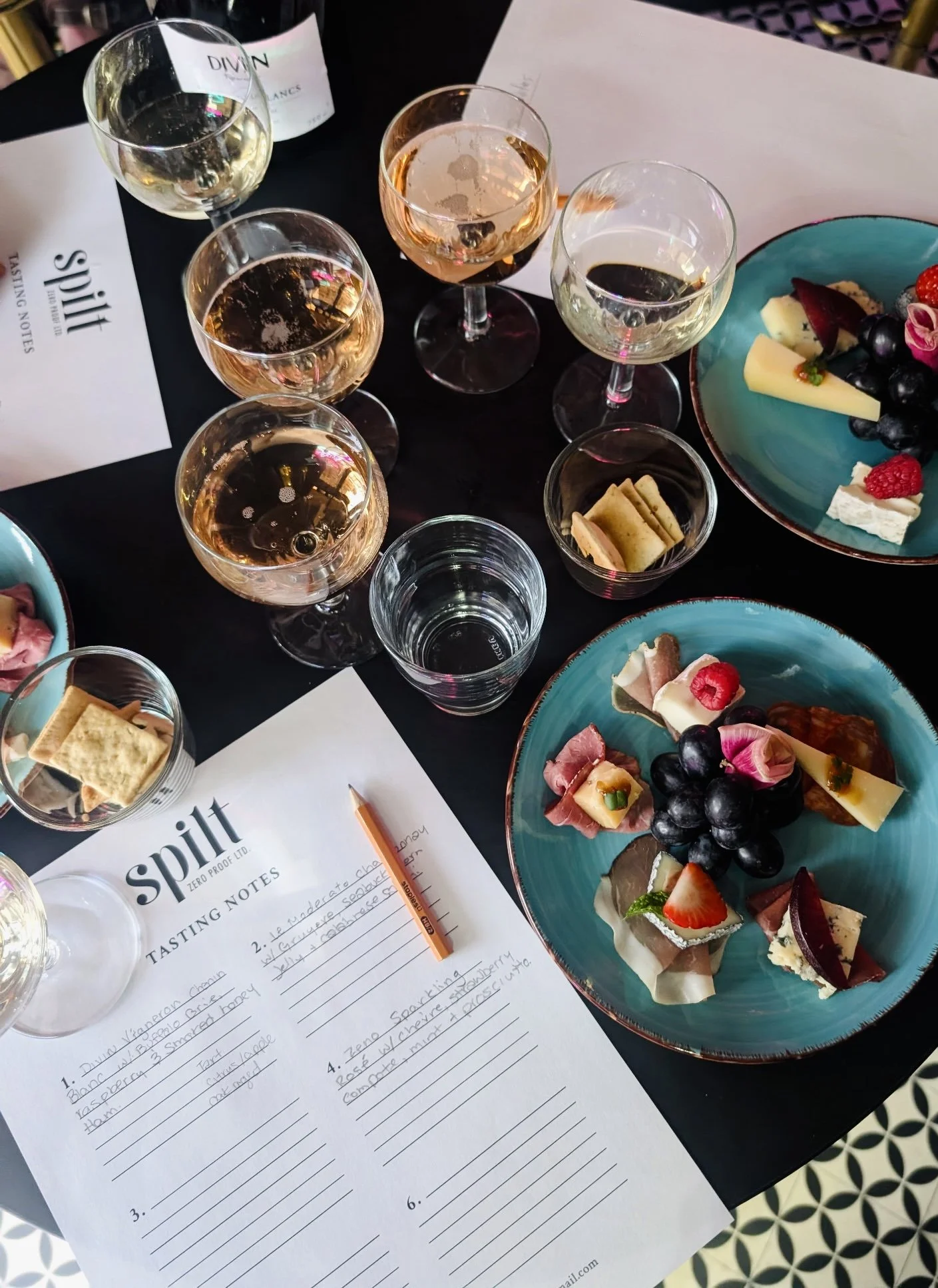 A tasting table set with several glasses of wine, a wine bottle, plates of assorted cheeses, fruits, and crackers, and tasting notes sheets with pencils.