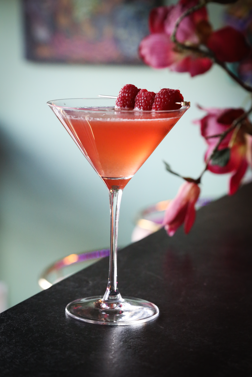 A pink cocktail garnished with three raspberries, served in a martini glass on a dark surface, with a blurred background of pink flowers and a painting.
