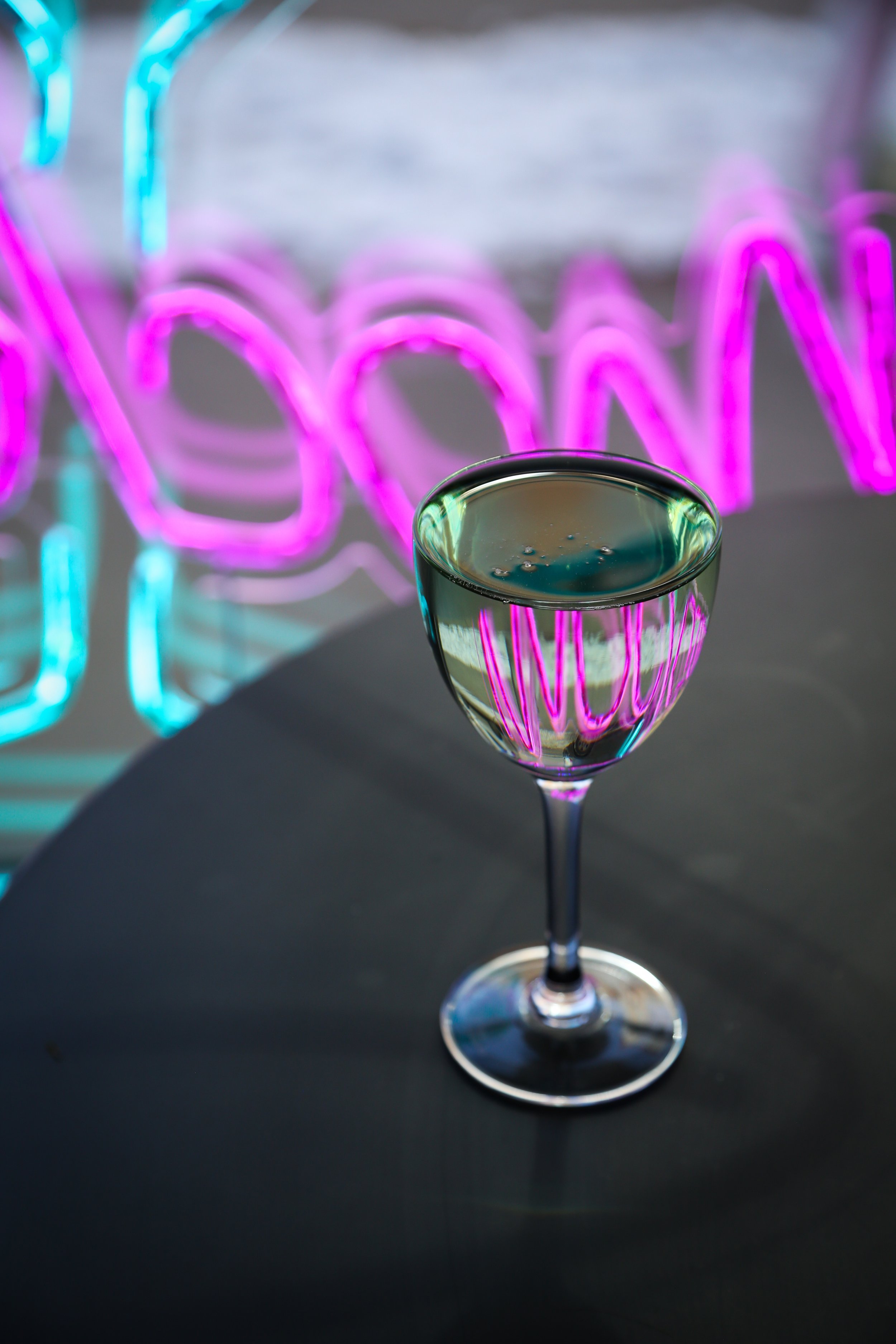 A glass of clear white liquid on a black surface with colorful neon light reflections.