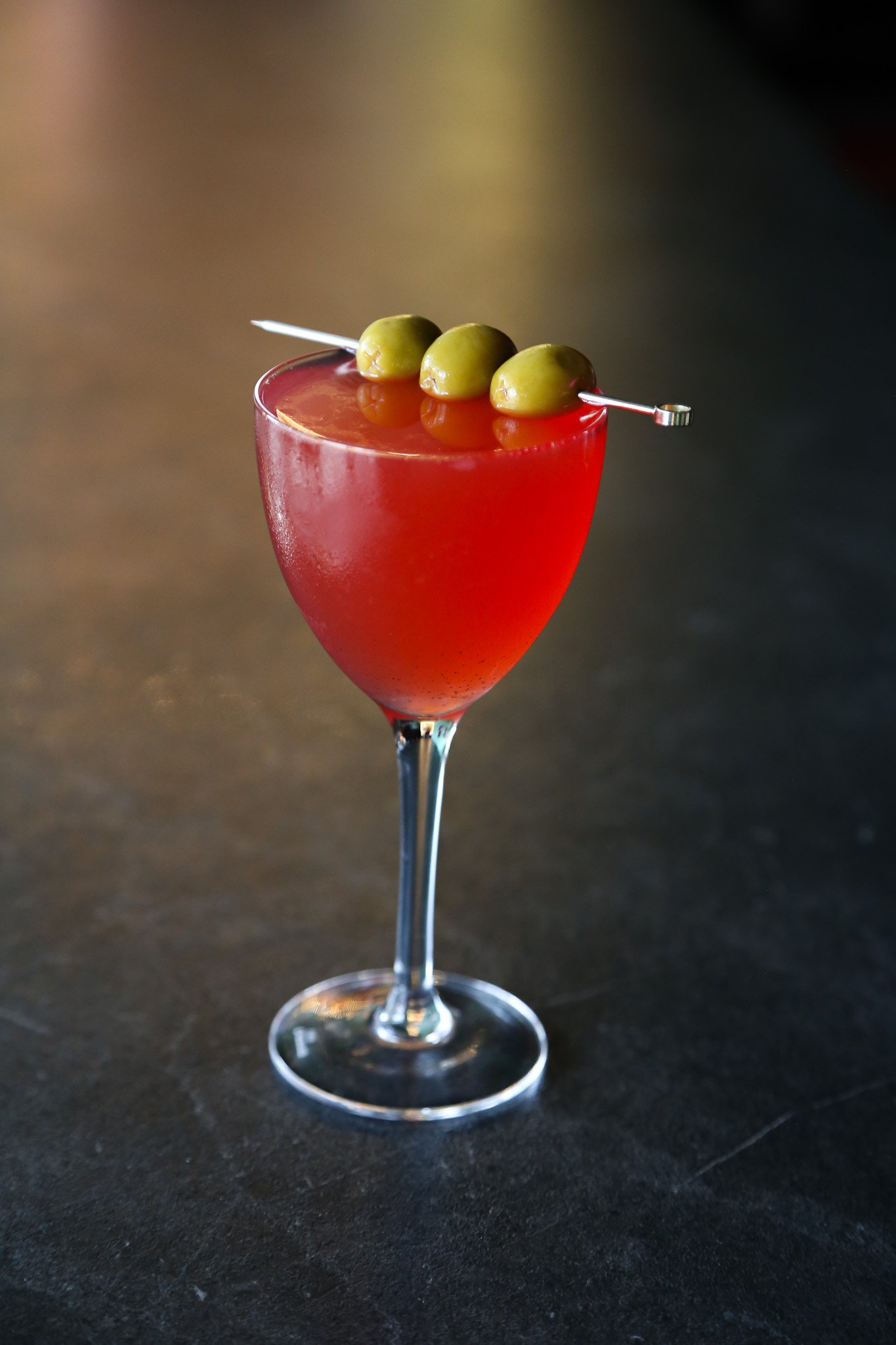 A red cocktail in a martini glass garnished with three green olives on a skewer against a dark background.