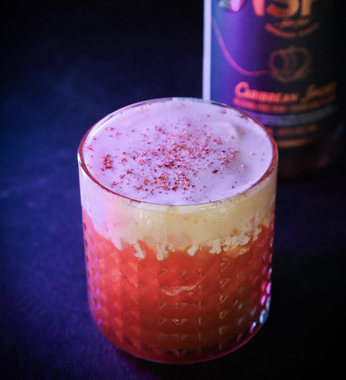 A layered cocktail in a textured glass with pink and yellow layers, topped with pink sprinkles, sitting on a dark surface with a can in the background.