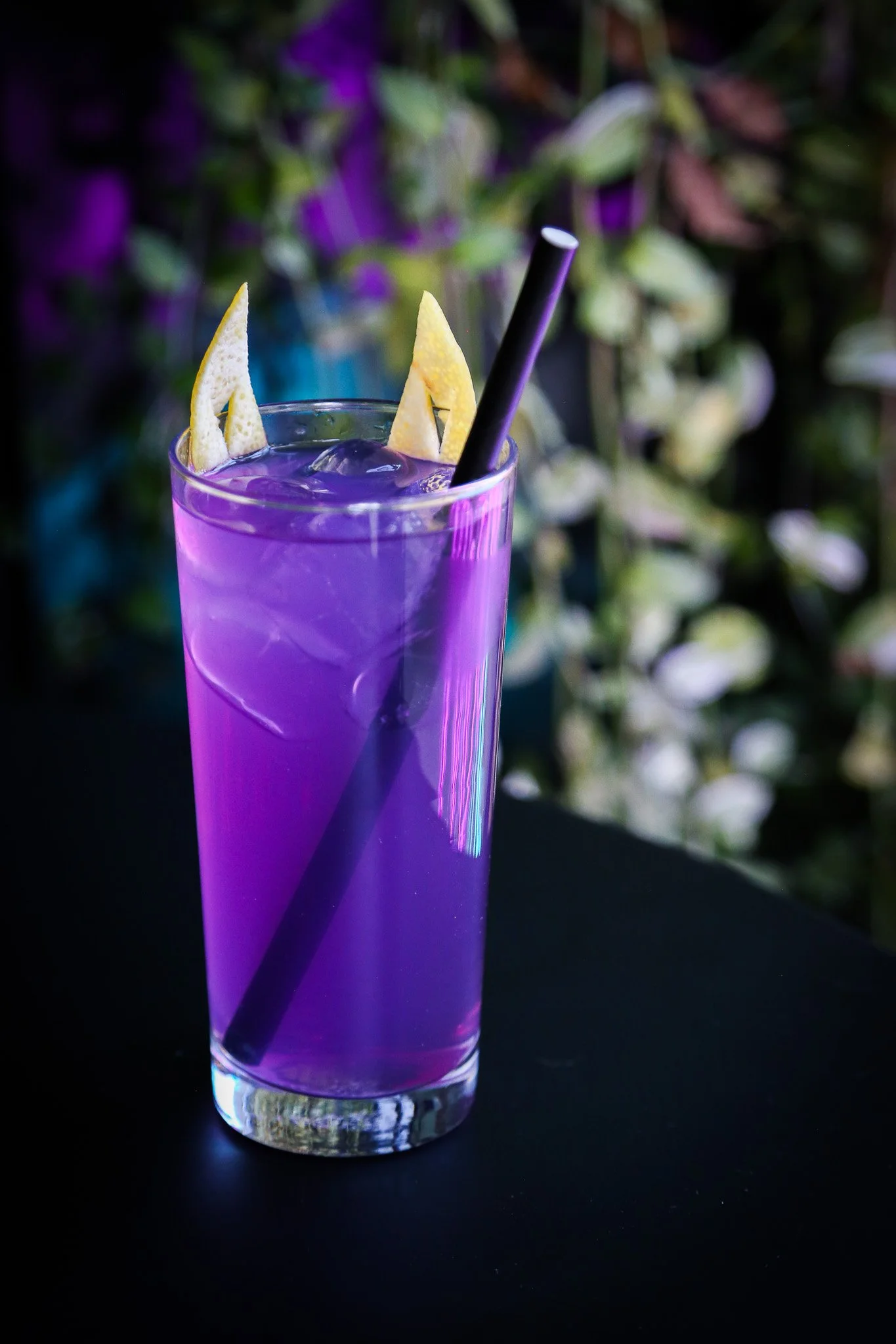 A tall glass filled with a purple beverage, ice cubes, and garnished with lemon wedges. There is a black straw in the drink, and the background features blurry greenery with purple and white flowers.