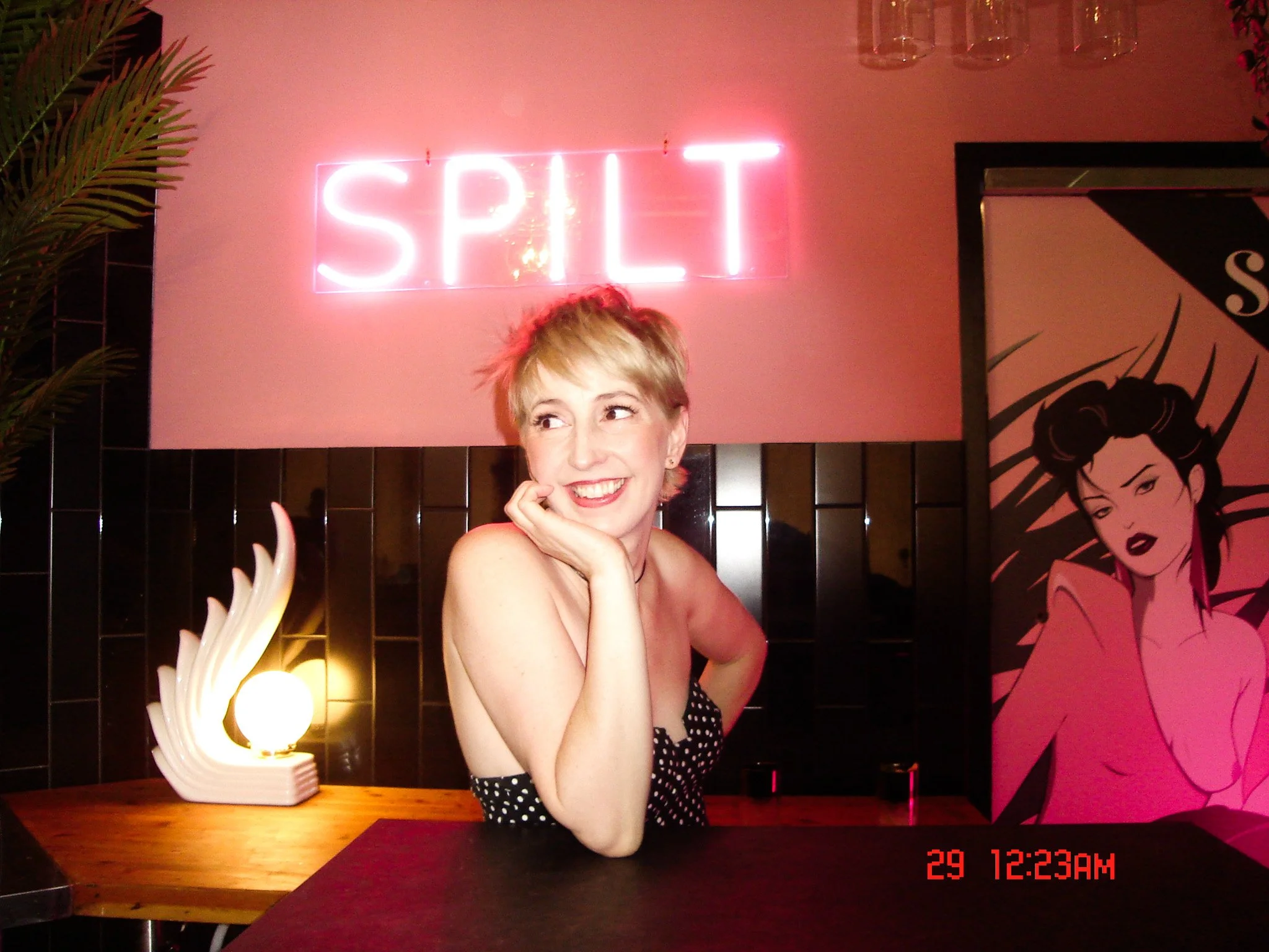 JoAnne Pearce with short blonde hair smiling and resting her chin on her hand, sitting at a table in a restaurant with pink walls and a neon sign that says 'SPILT' behind her.
