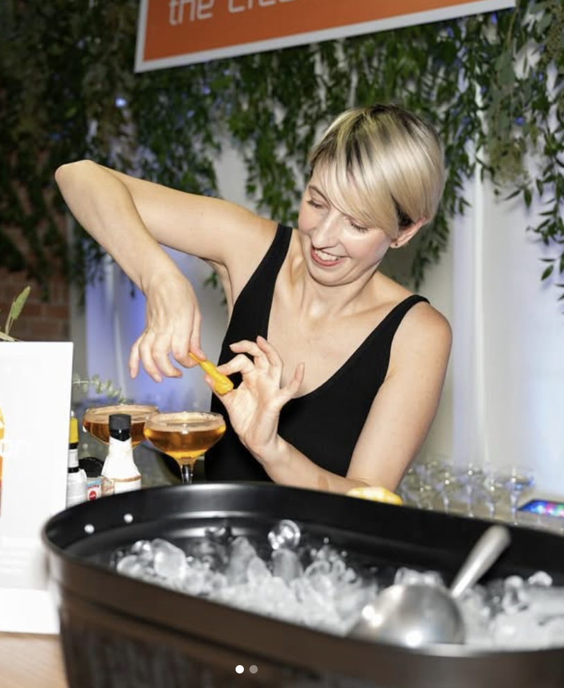 A woman with short blonde hair and wearing a black tank top is smiling and preparing drinks at a bar or event table. She is squeezing a yellow citrus juicer over two cocktail glasses filled with a light brown beverage. A large black container of ice with a metal scoop is in the foreground, and there are small bottles and decorations on the table. Green foliage and a partially visible sign are in the background.