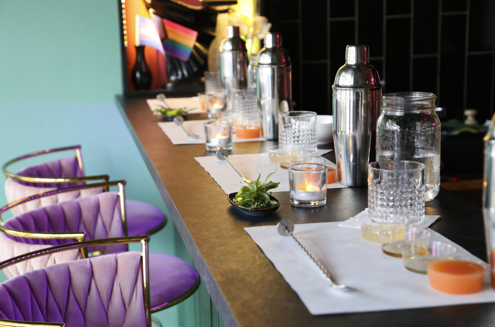 A decorated bar or dining counter with glasses, bottles, candles, and small garnish plates, with pink chairs and a teal wall in the background.