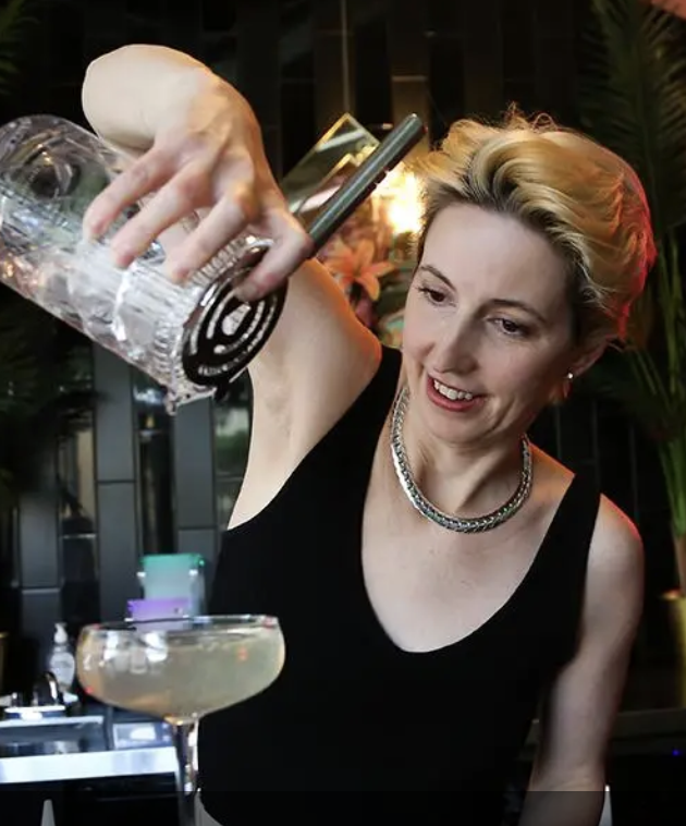 JoAnne Pearce pours a clear liquid from a large bottle into a cocktail glass at a bar or restaurant, with a smile on her face.