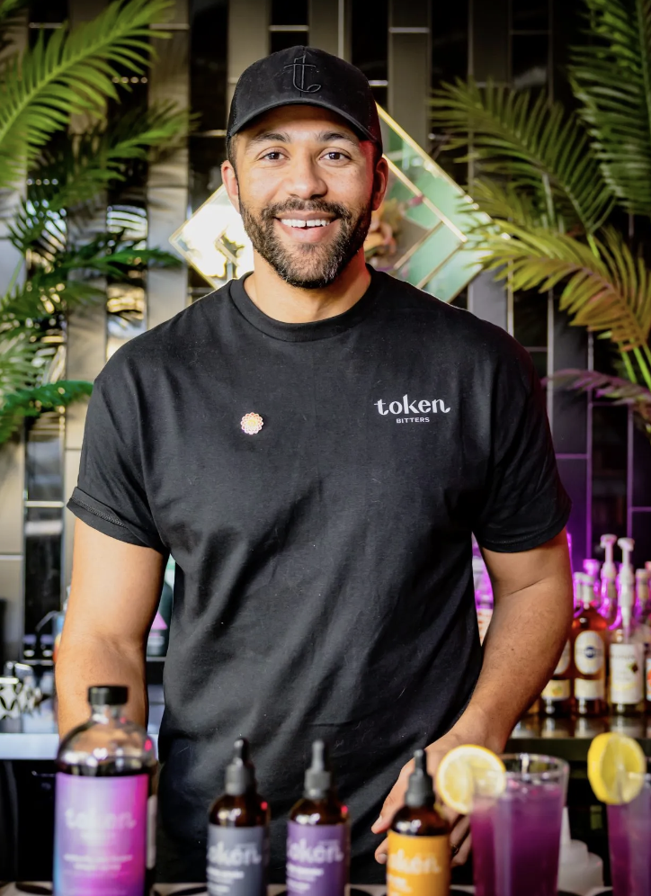 Keenan Pasal in a black t-shirt and cap standing behind a bar with colorful bottles and drinks, surrounded by green plants.