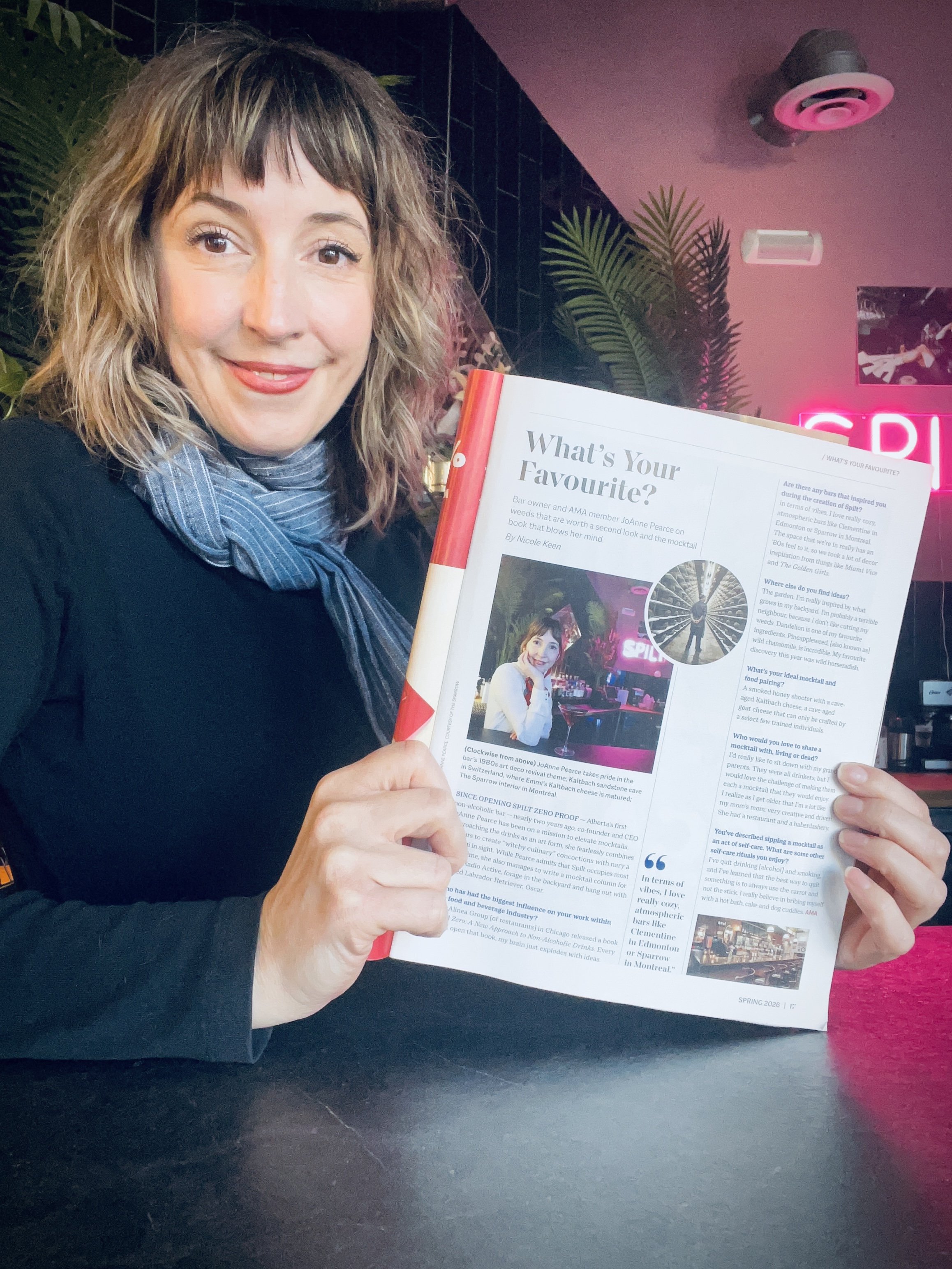 JoAnne Pearce holds an open magazine titled 'What's Your Favourite?' in a dimly lit room with tropical decor and a pink neon sign.
