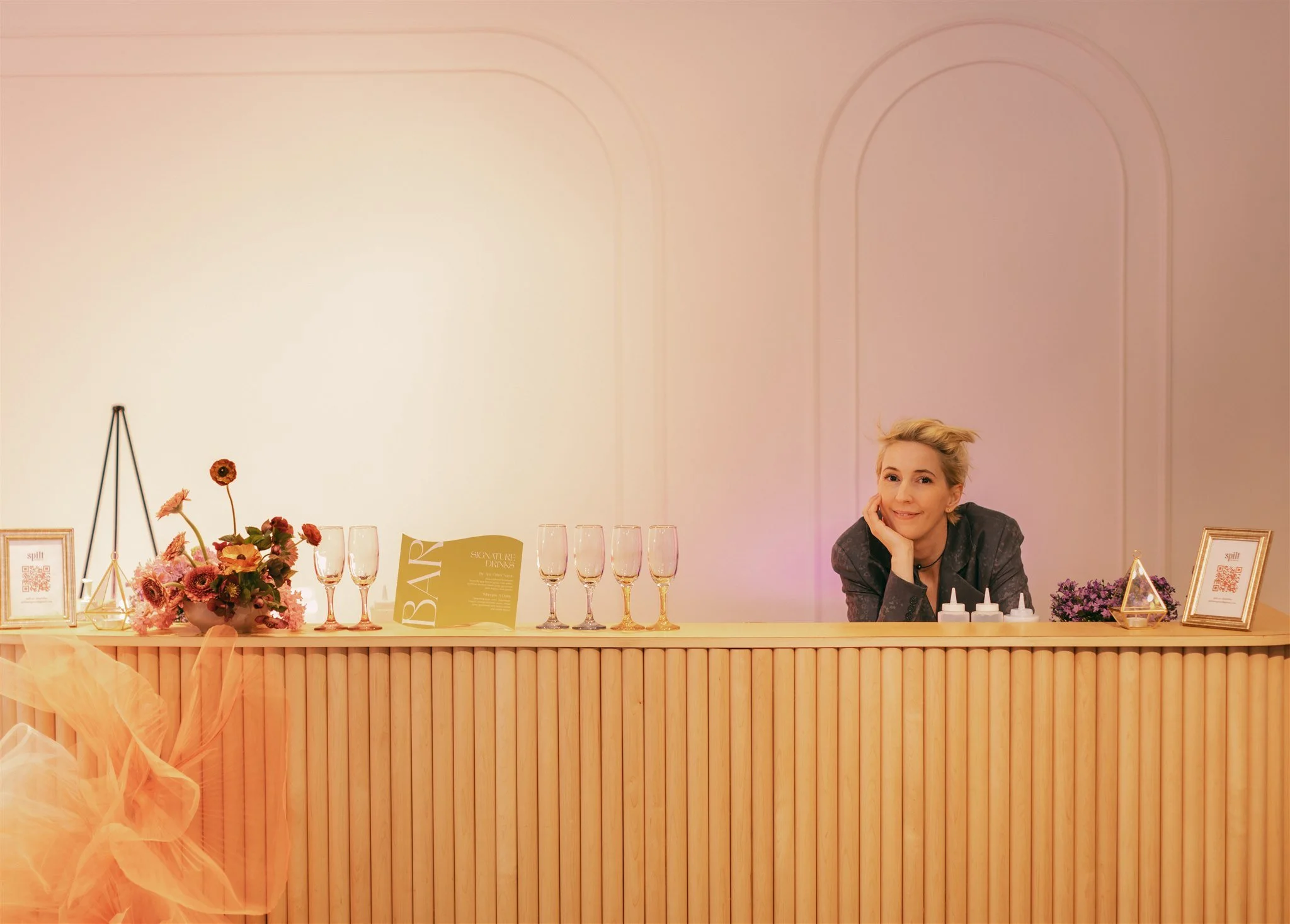 JoAnne Pearce sitting at a wedding or event reception table decorated with pink flowers, framed signs, glasses, and bottles. The table has a wooden base and the background is a light pink wall with decorative arches.