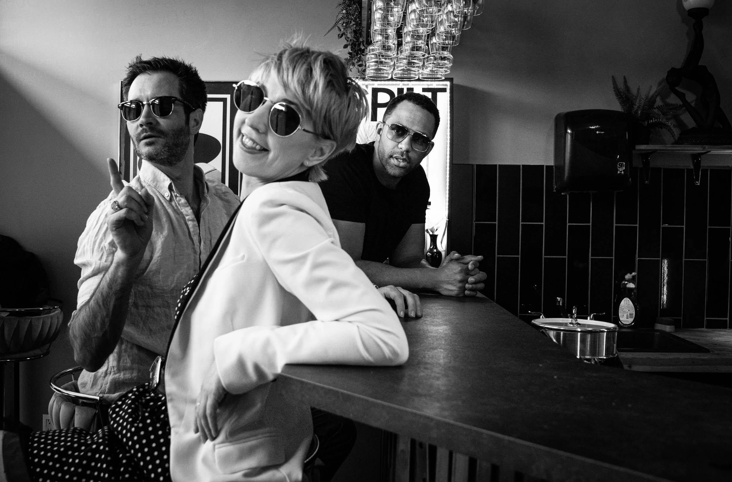 Black and white photo of three people sitting at a bar; two men and one woman, all wearing sunglasses. The woman is smiling and appears to be talking or laughing. The man on the left is pointing upward with his index finger. The man on the right is looking towards the camera. The background includes a sign, glasses hanging upside down, and some plants.