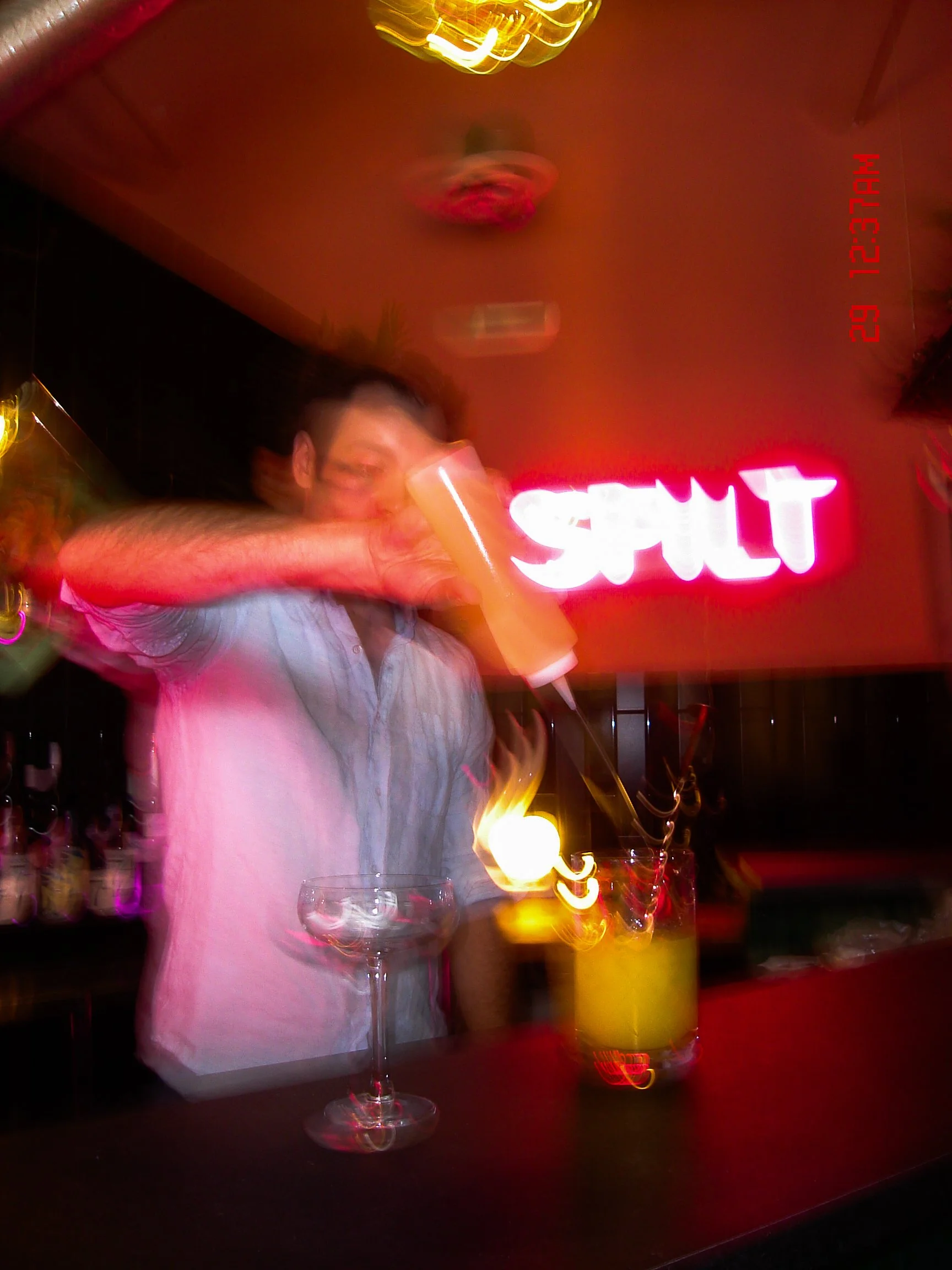 Andrew Paul holding a drink in a glass at a bar, with bottles of alcohol in the background and a neon sign that says 'craft'. The setting appears to be a dimly lit bar or nightclub.