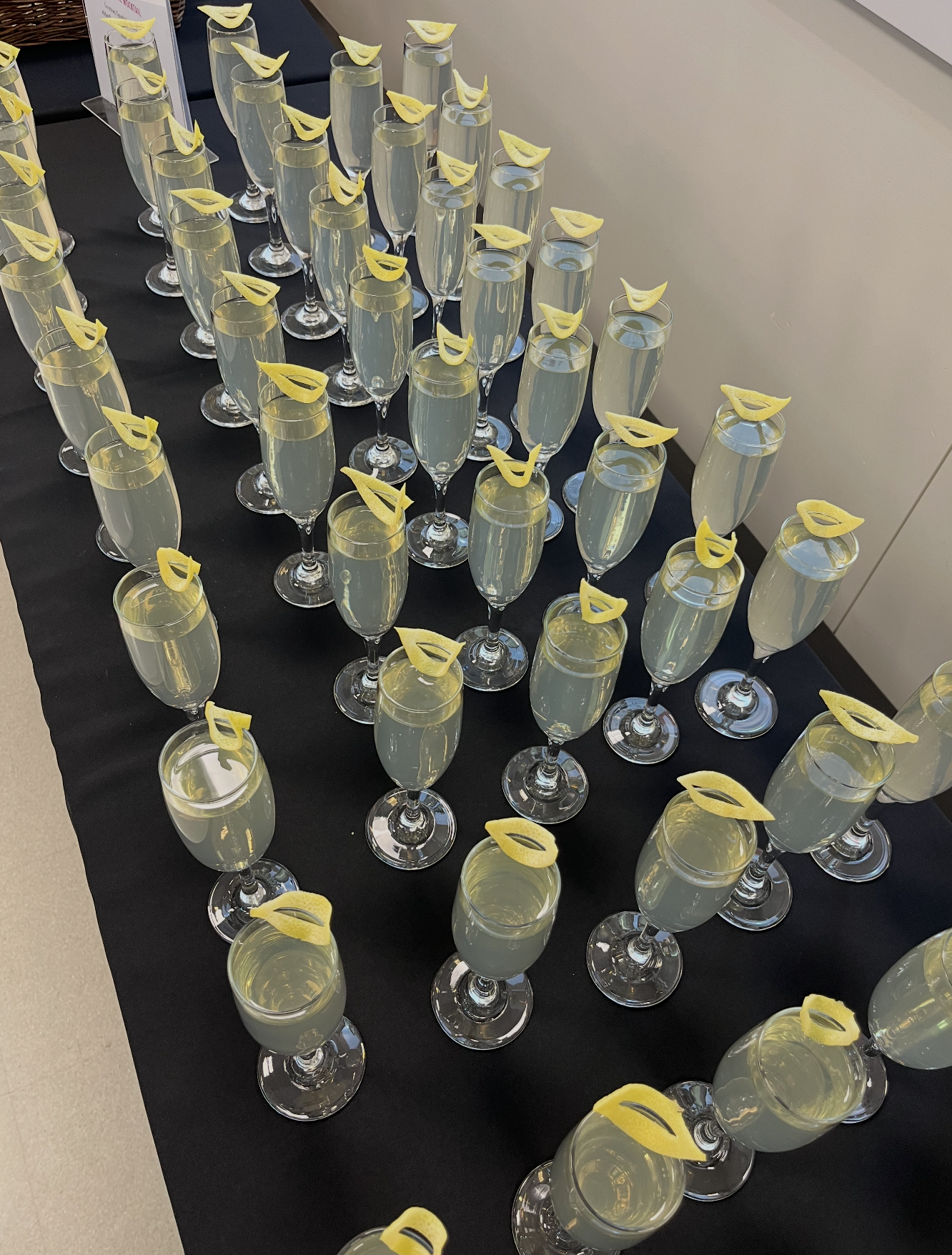 Multiple champagne flutes filled with a light yellow beverage, each garnished with a lemon twist, arranged on a black tablecloth.
