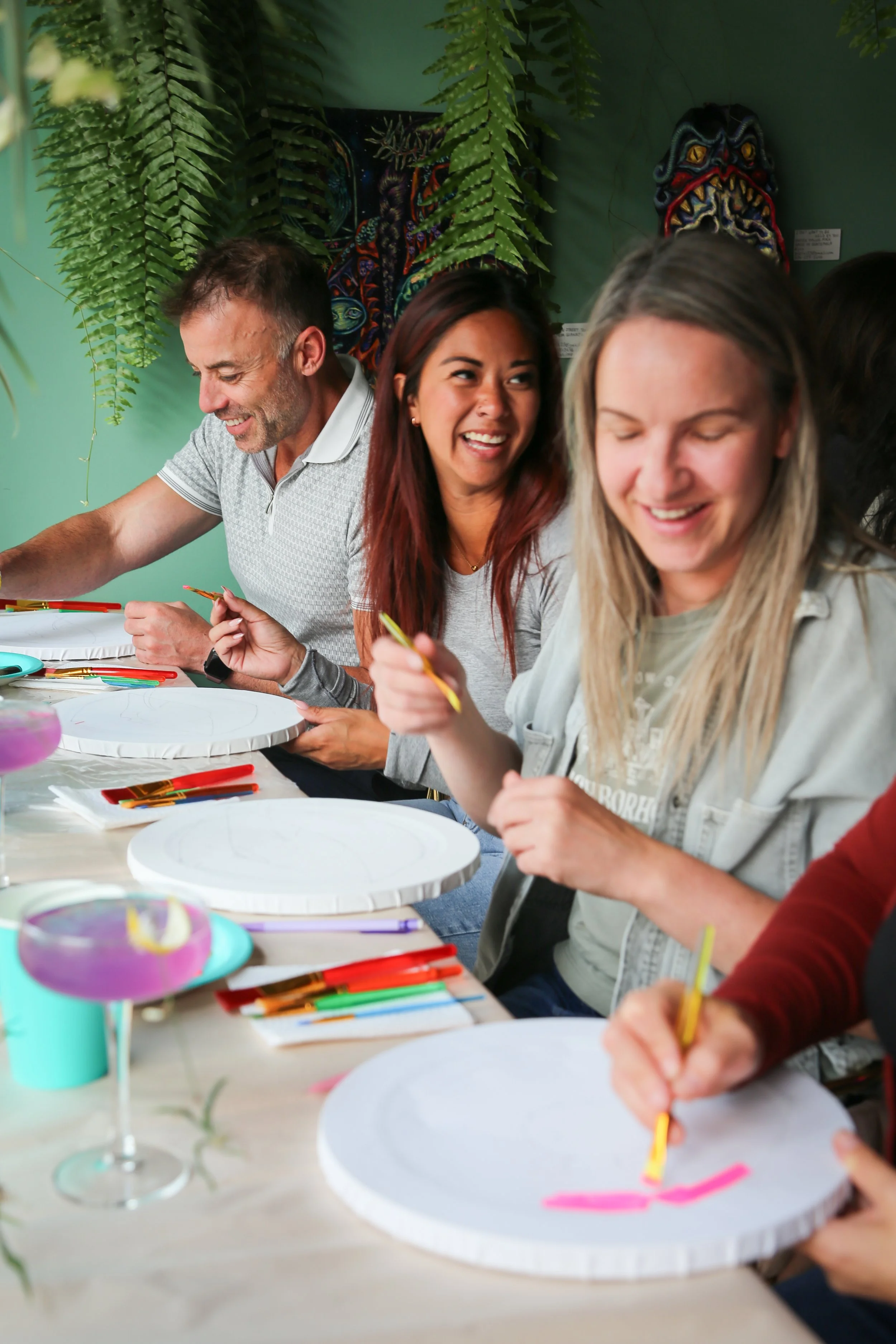 A group of people sitting at a table engaging in a paint night, surrounded by colorful supplies and artwork on the wall.