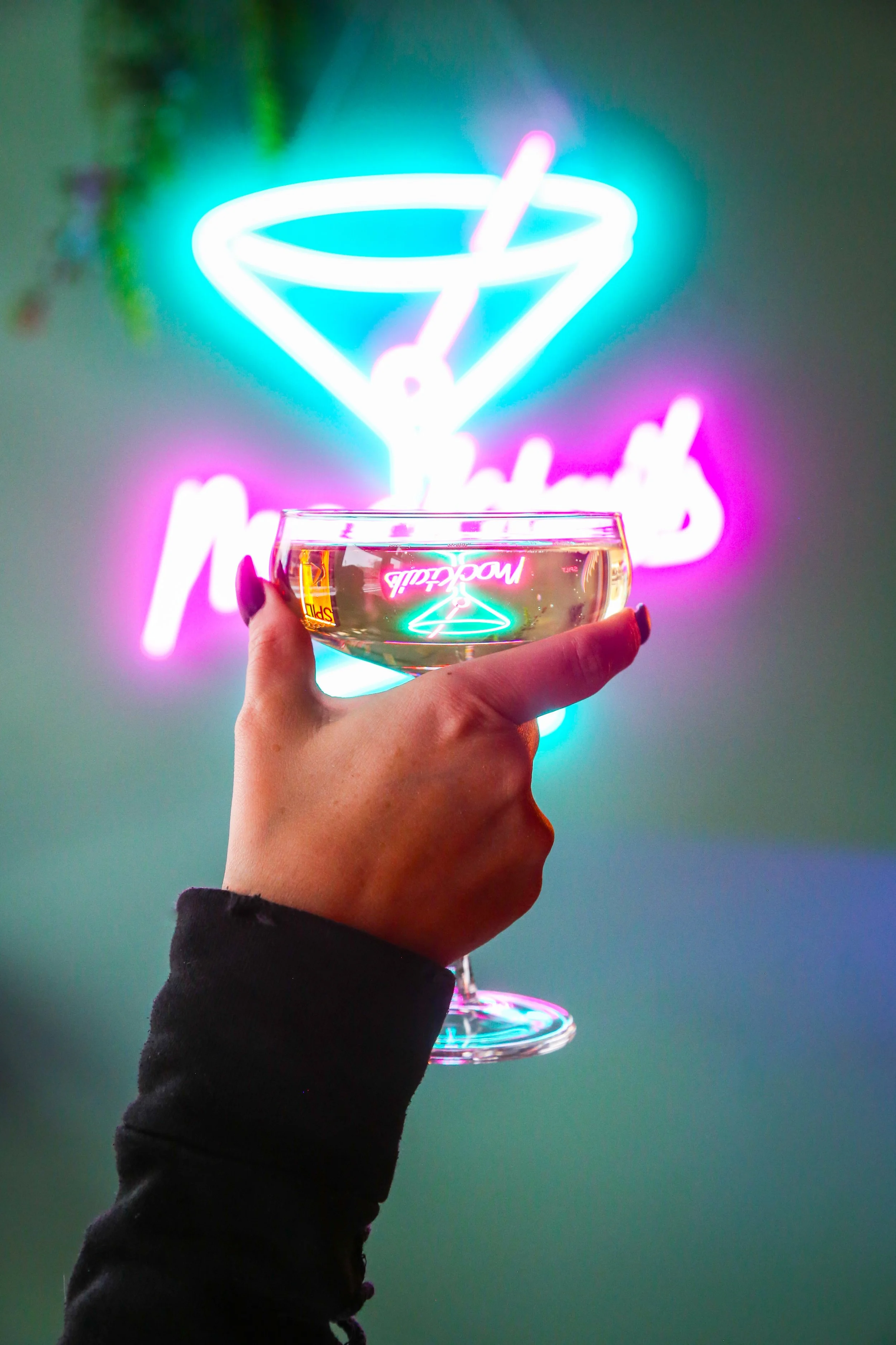 A hand holding a champagne coupe glass filled with a clear cocktail, with a neon sign of a martini glass in the background and the word 'Mocktail' glowing in pink and turquoise.