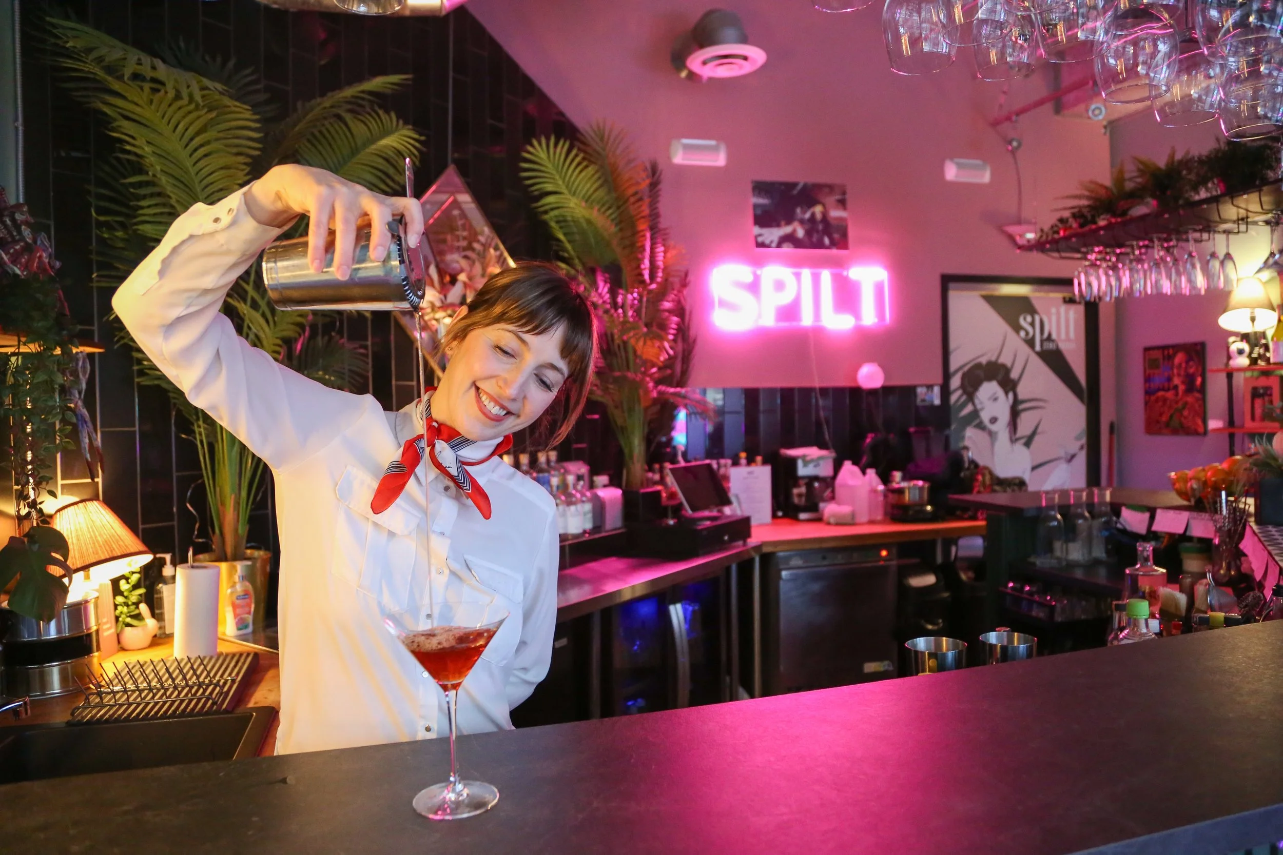 JoAnne Pearce in a white blouse and jacket and red neck scarf smiles as she pours a drink into a martini glass at a bar with pink neon lighting and green plants. Photo for AMA Insider.