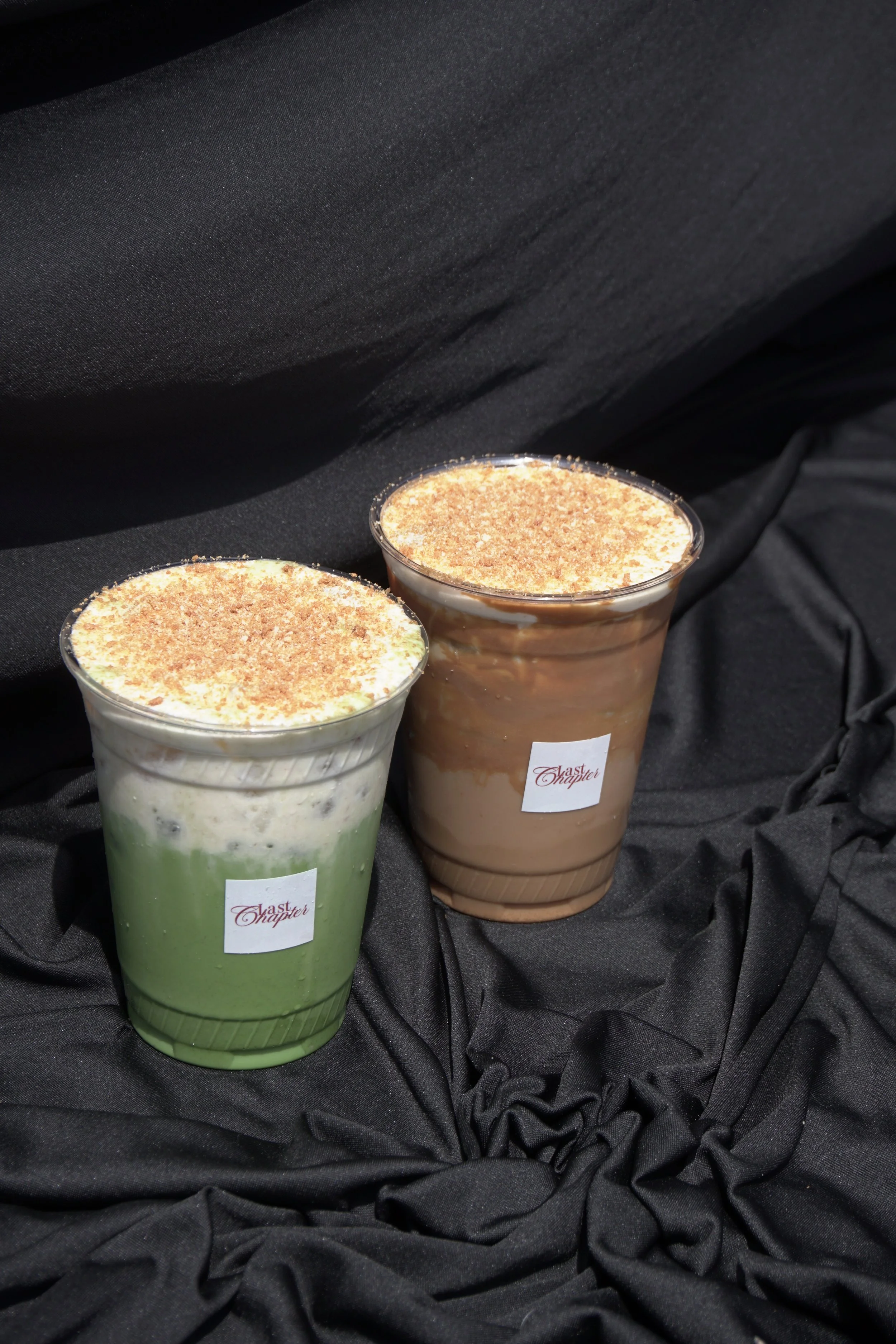 Two iced beverages in clear plastic cups with brown and green layers, topped with powder, on black fabric background.