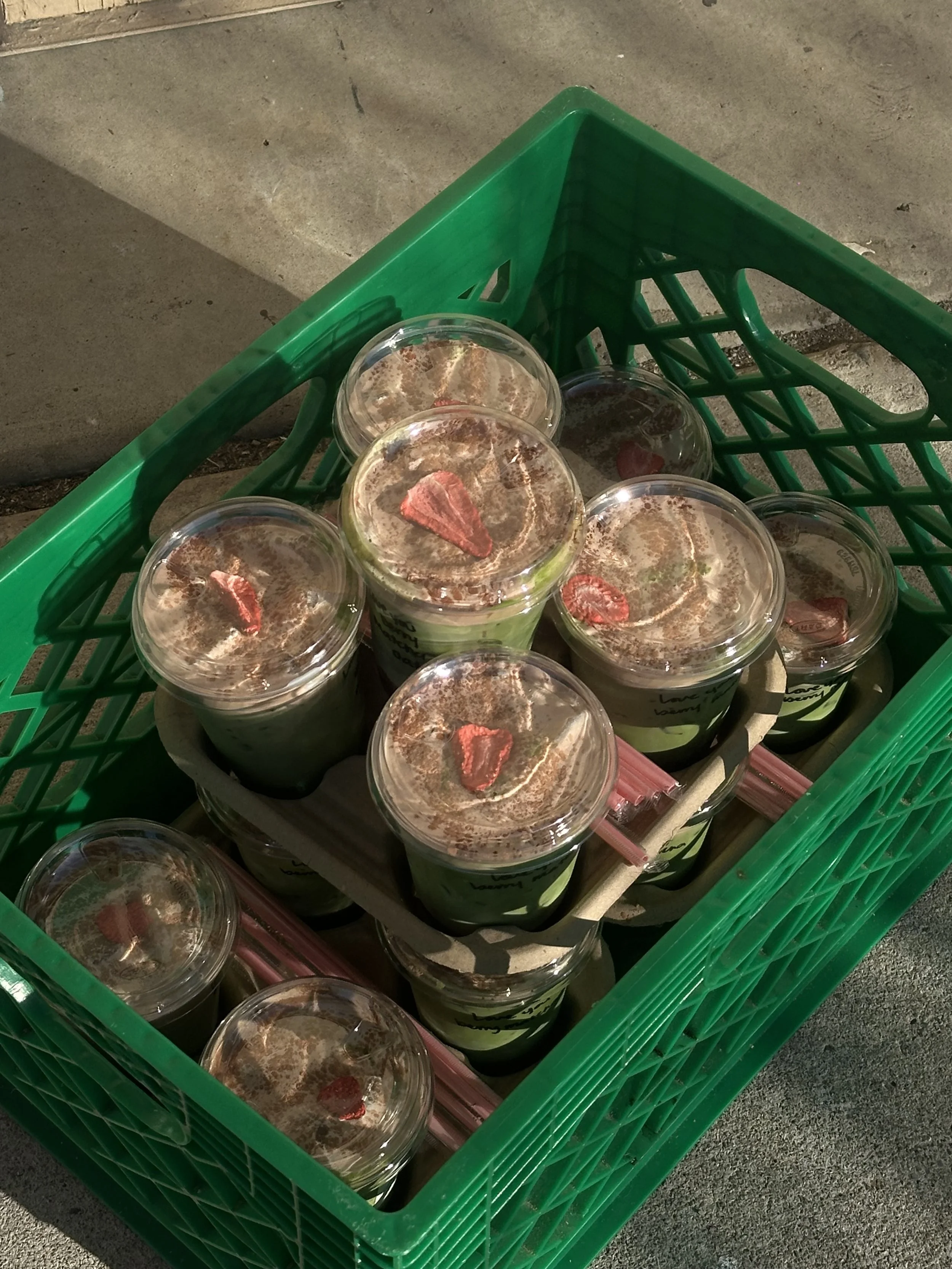 Green shopping basket filled with several cups of chocolate or coffee drinks topped with whipped cream and a small piece of candy or fruit on top, sitting on a sidewalk. Bay Area coffee and matcha catering for corporate events, brand activations, wed